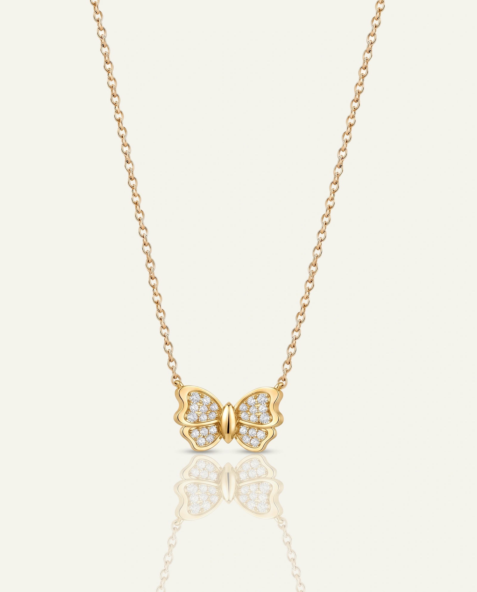 A delicate gold chain necklace featuring a butterfly pendant paved with small clear stones, centered on a light cream background with a soft reflection below.