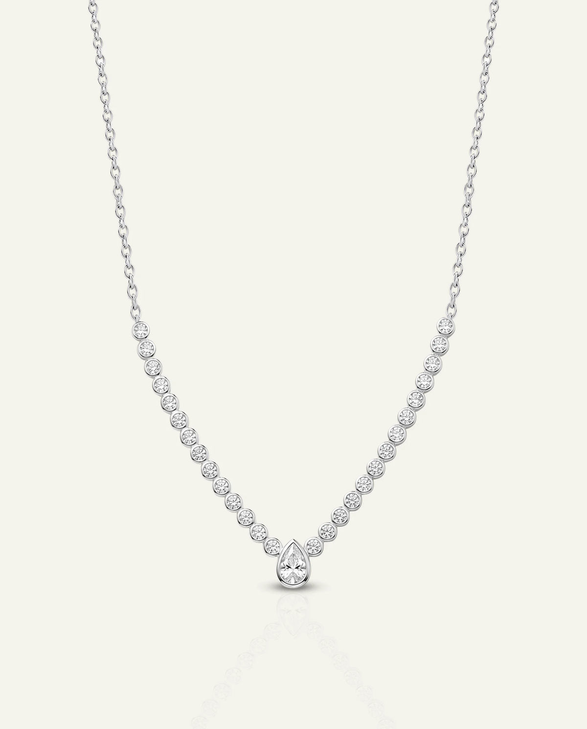 A delicate silver tennis necklace with a pear-cut diamond pendant, displayed on a light cream background.