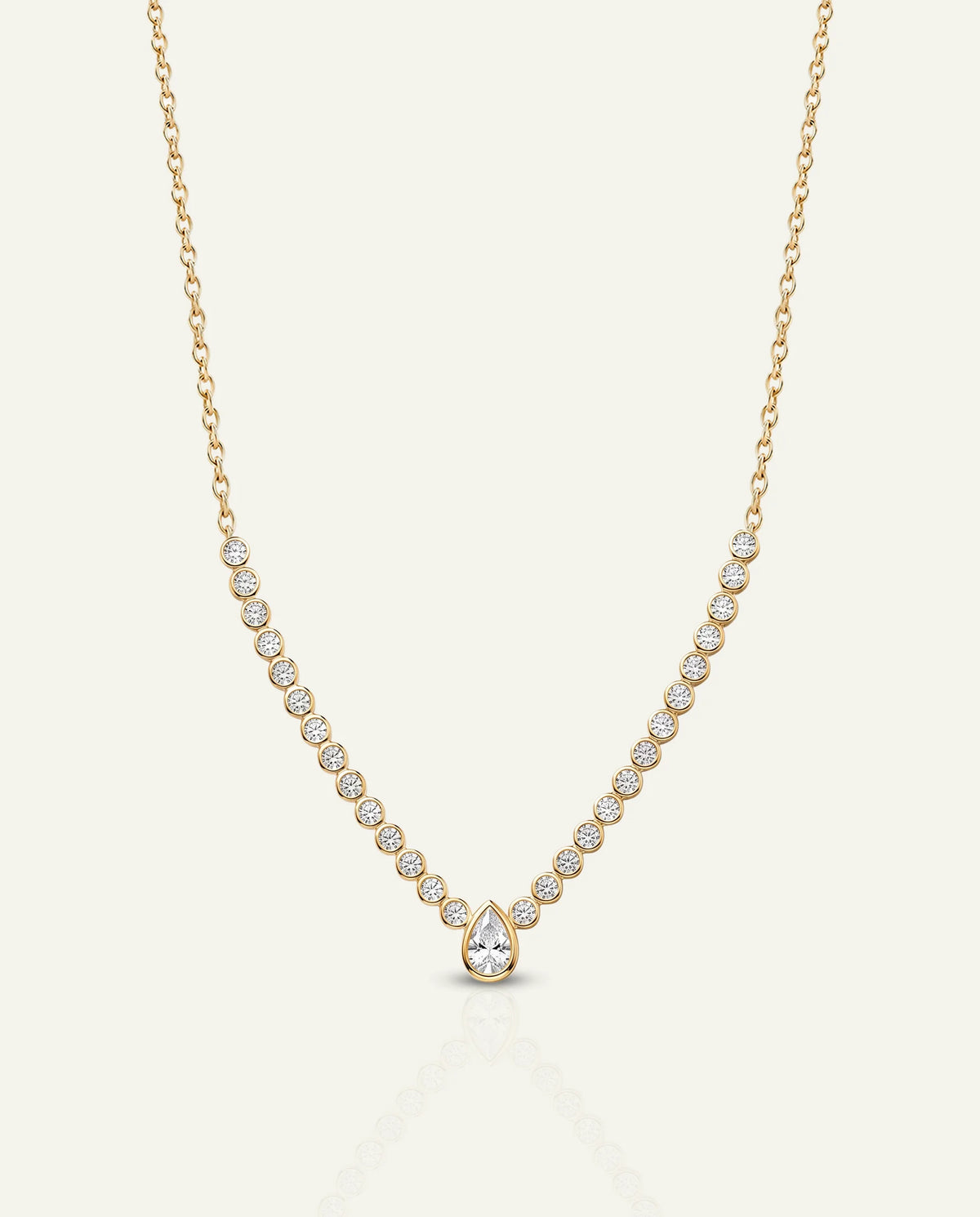 A delicate gold necklace featuring a line of small round-cut clear stones leading to a pear-shaped clear stone pendant, displayed on a light cream background.