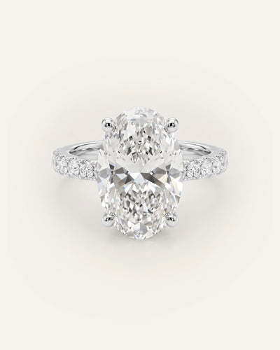 An oval-cut diamond engagement ring with a silver-toned band set with small pavé diamonds, centered on a light cream background.