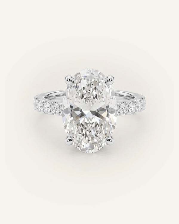 An oval-cut diamond engagement ring with a silver-toned band set with small pavé diamonds, centered on a light cream background.