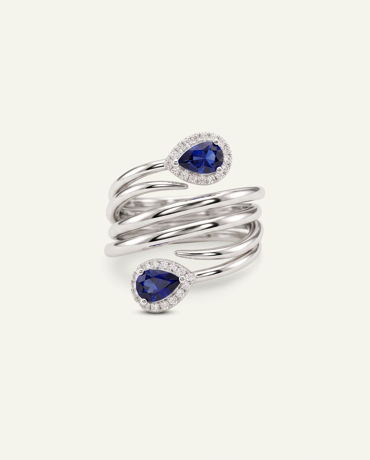A silver wrap ring featuring two pear-cut blue sapphires, each surrounded by a halo of small clear stones, set against a solid off-white background.