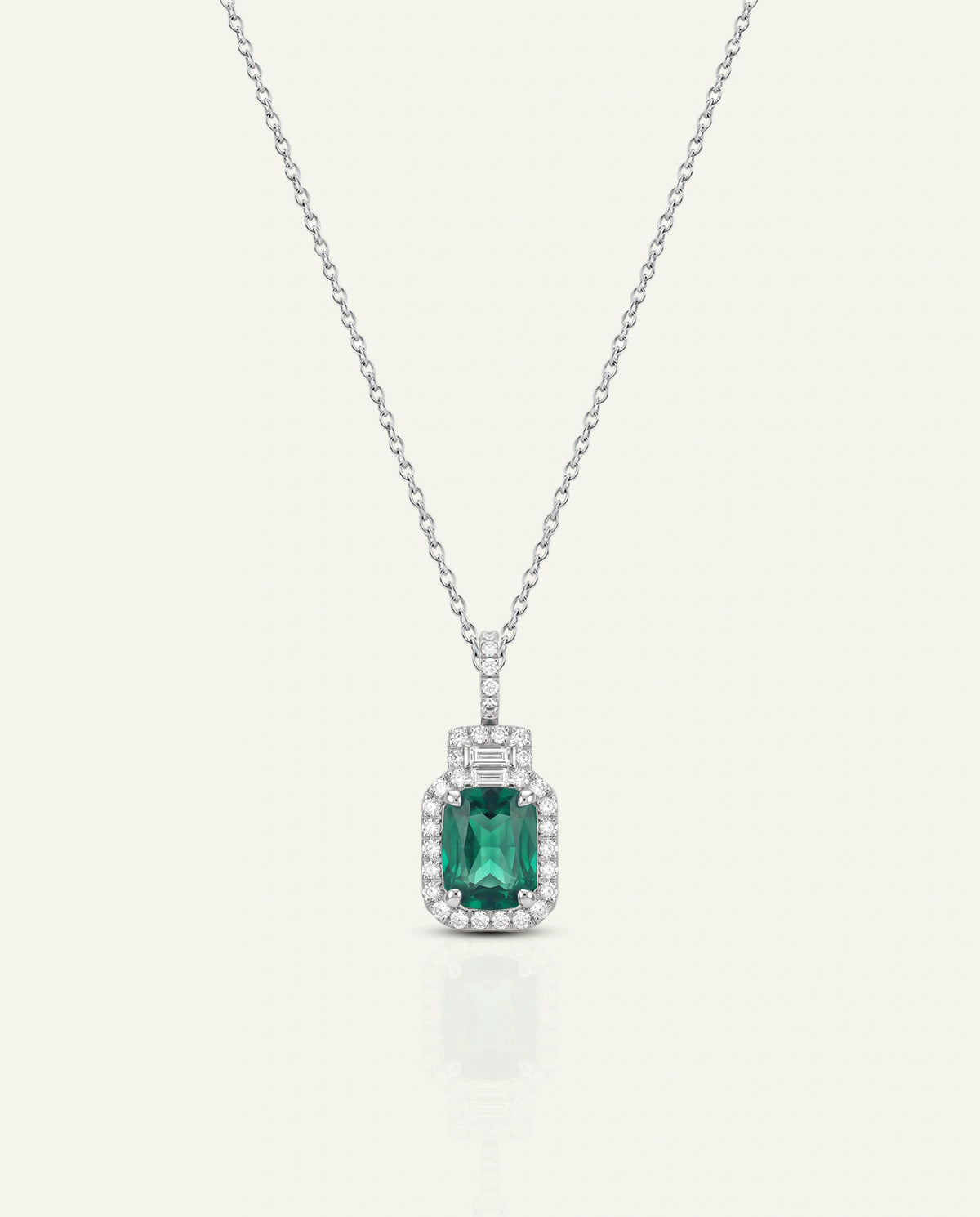 A silver pendant necklace featuring a rectangular green emerald-cut center stone surrounded by a halo of small clear stones, suspended from a delicate silver chain on a light cream background.