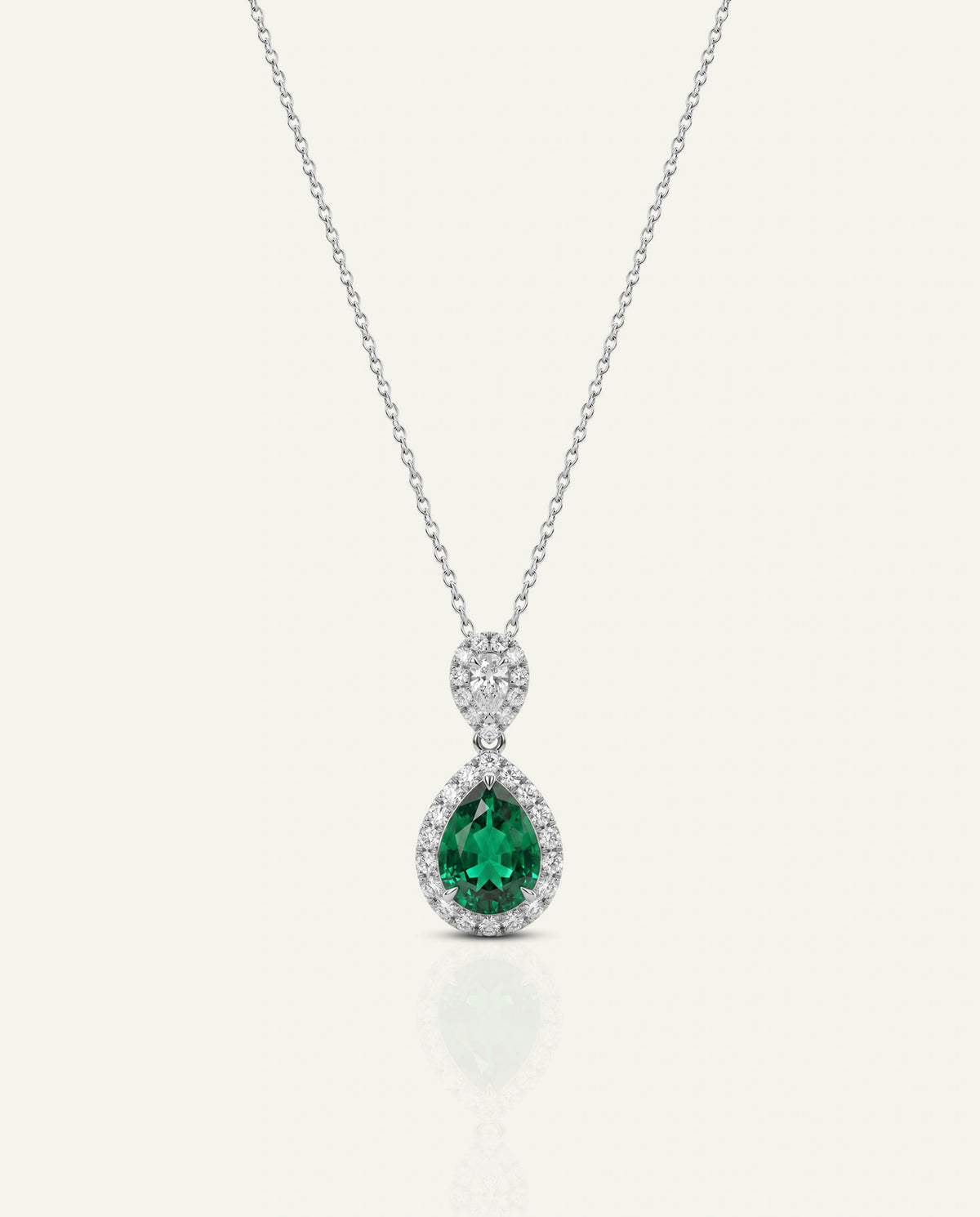 A pear-cut emerald green gemstone pendant necklace in a silver-toned setting, surrounded by small clear stones, hanging from a delicate silver chain against a light cream background.