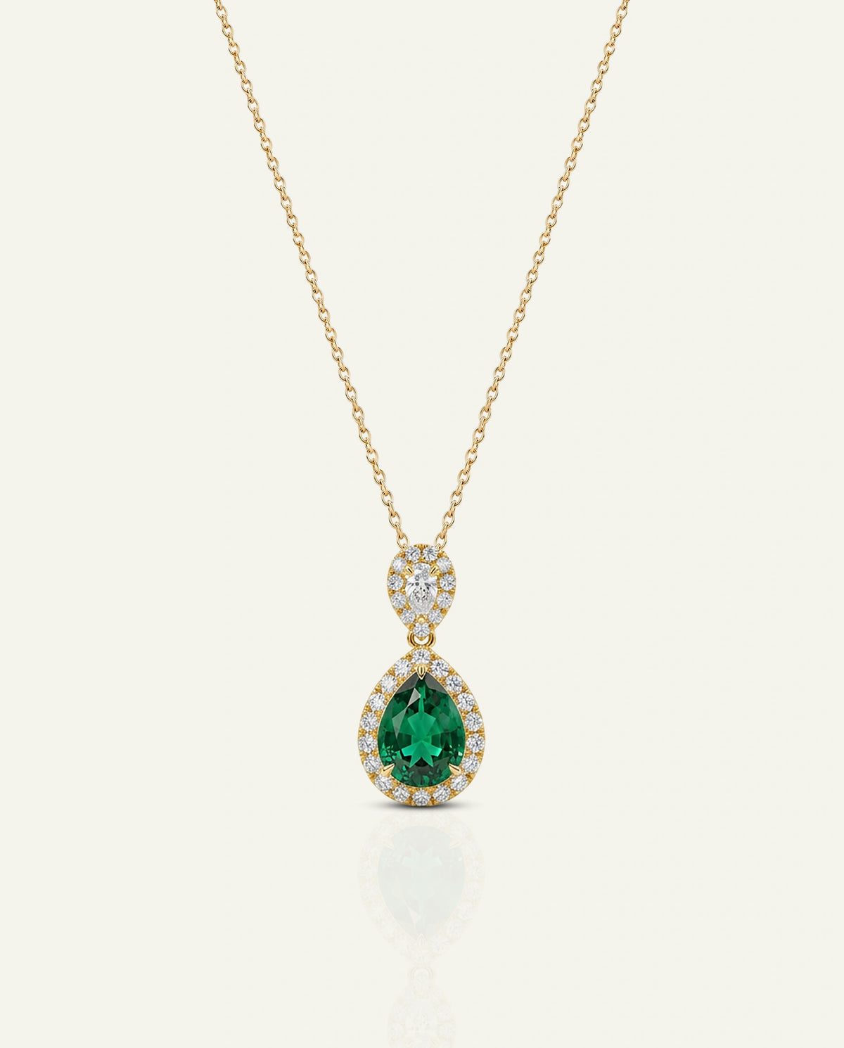 A gold pendant necklace featuring a teardrop-shaped emerald-cut green gemstone surrounded by small clear stones, suspended from a delicate gold chain on a light cream background.