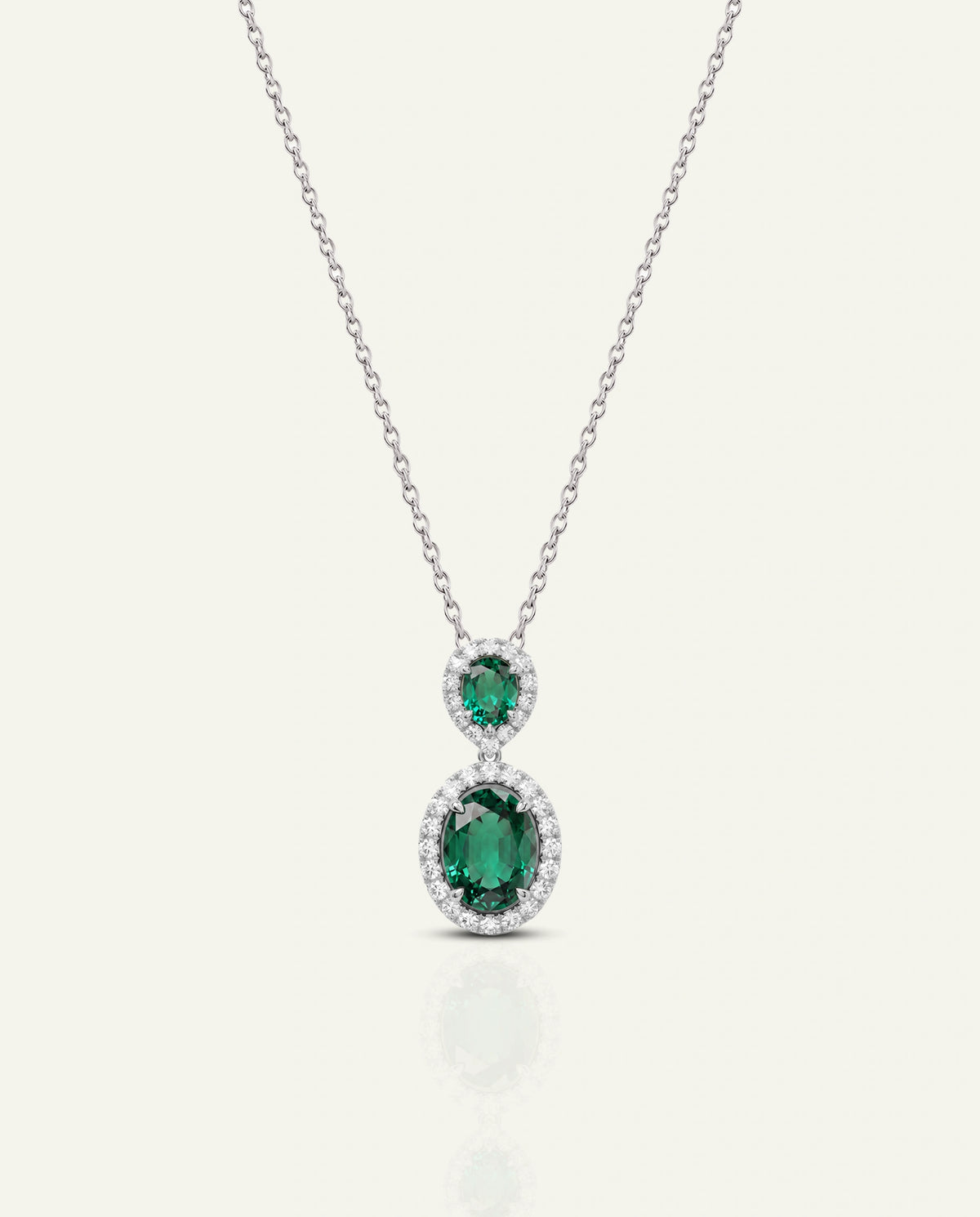 A silver-tone pendant necklace featuring two emerald-cut green gemstones surrounded by small clear stones, suspended from a delicate chain on a light cream background.