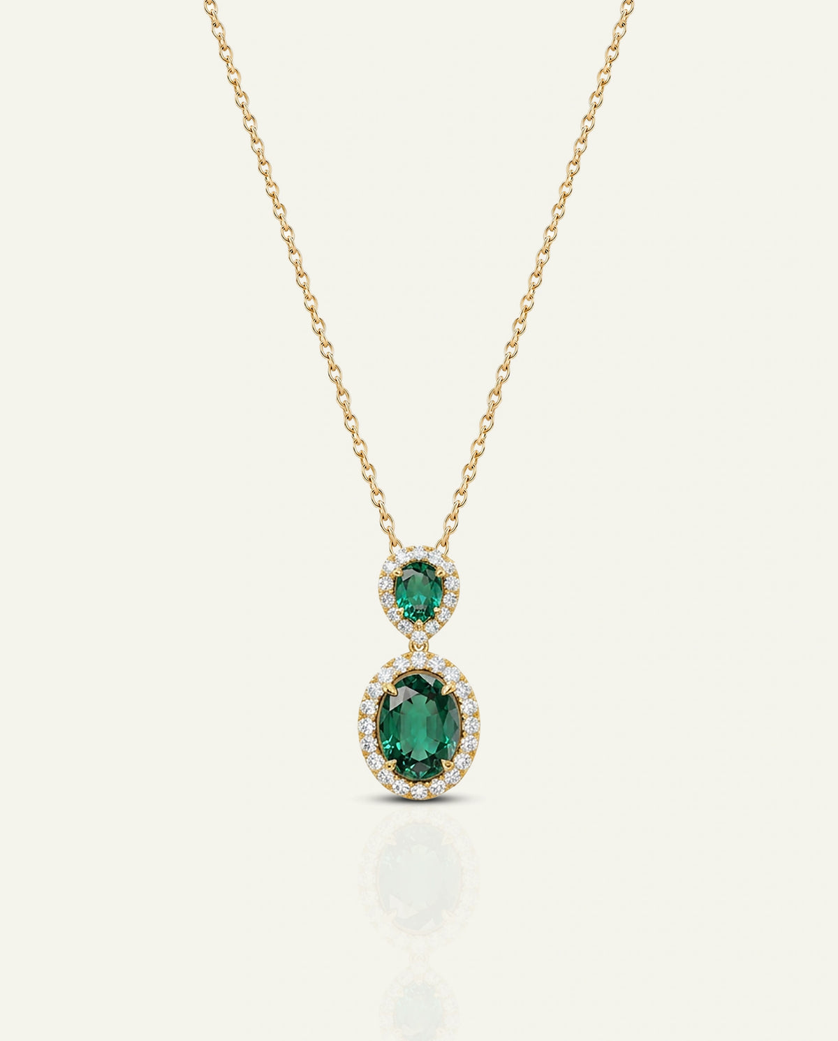 A gold necklace featuring a two-stone pendant with a smaller oval-cut green gemstone and a larger oval-cut green gemstone, both surrounded by small clear stones, on a light cream background.