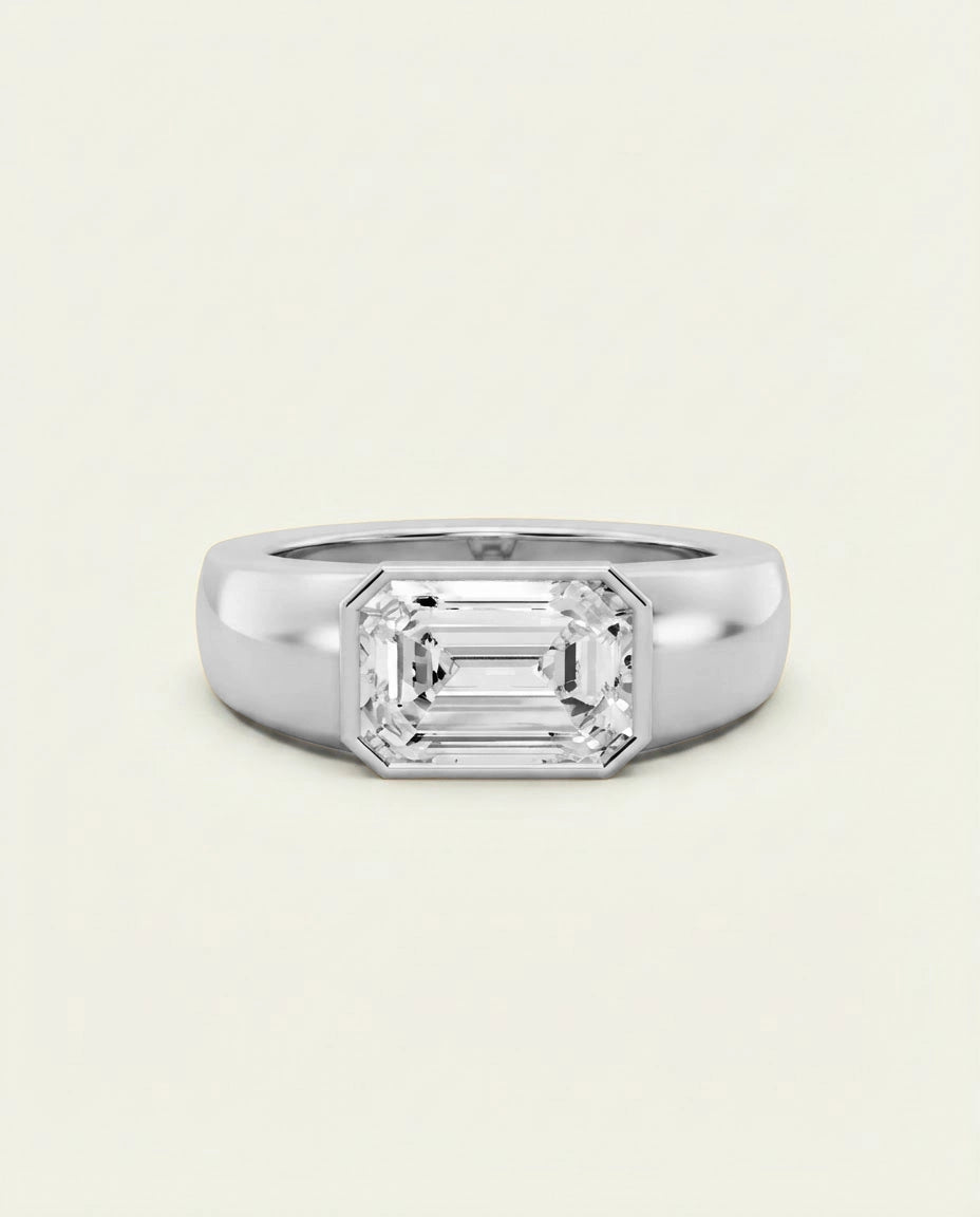 An emerald-cut diamond ring in a polished silver-colored metal band, centered on a light cream background.