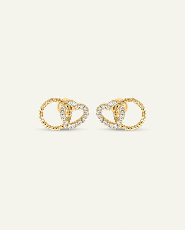 A pair of gold heart-and-ring stud earrings with small clear stones, shown on a plain light warm-gray background.