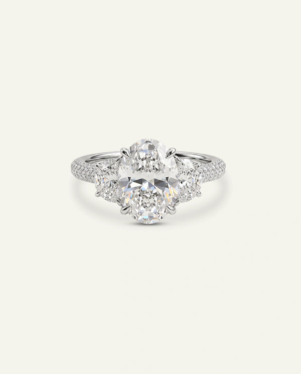 A silver-toned diamond ring with a central round brilliant-cut diamond surrounded by smaller round and oval diamonds, set on a delicate pavé band, shown against a solid off-white background.