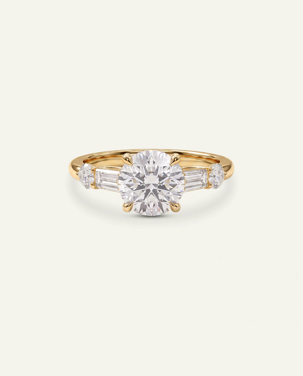 A yellow gold ring featuring a large round-cut center stone flanked by baguette and round side stones, set against a plain off-white background.