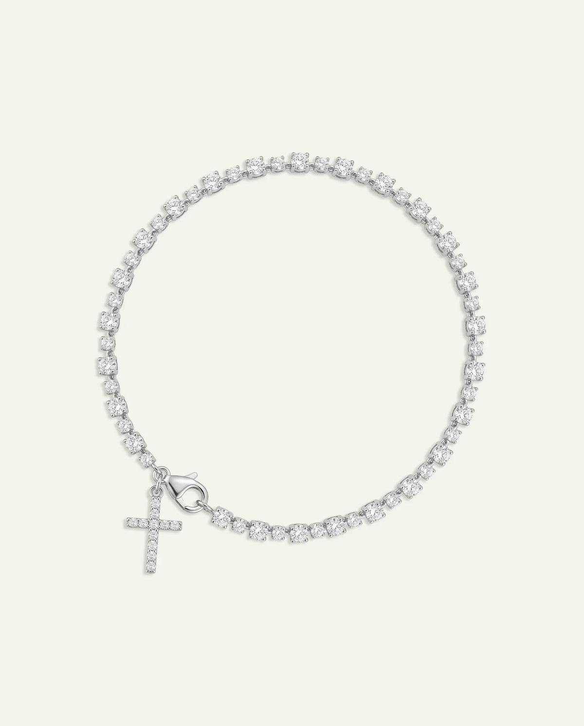 A delicate silver-toned tennis bracelet with a small cross charm, set with clear stones, displayed on a light cream background.