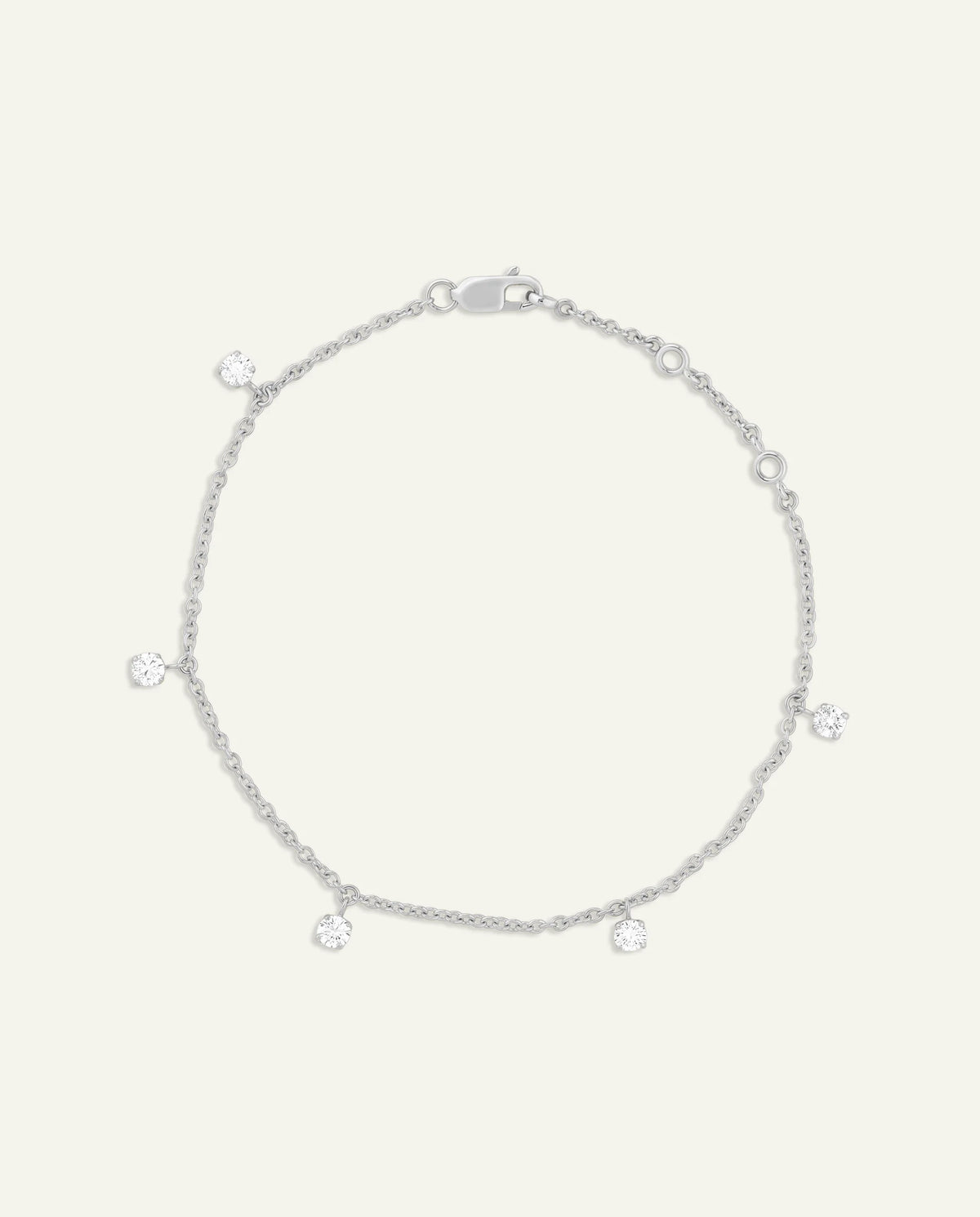 A delicate silver chain anklet with five small, clear round-cut gemstone charms spaced evenly along the chain, displayed on a light cream background.