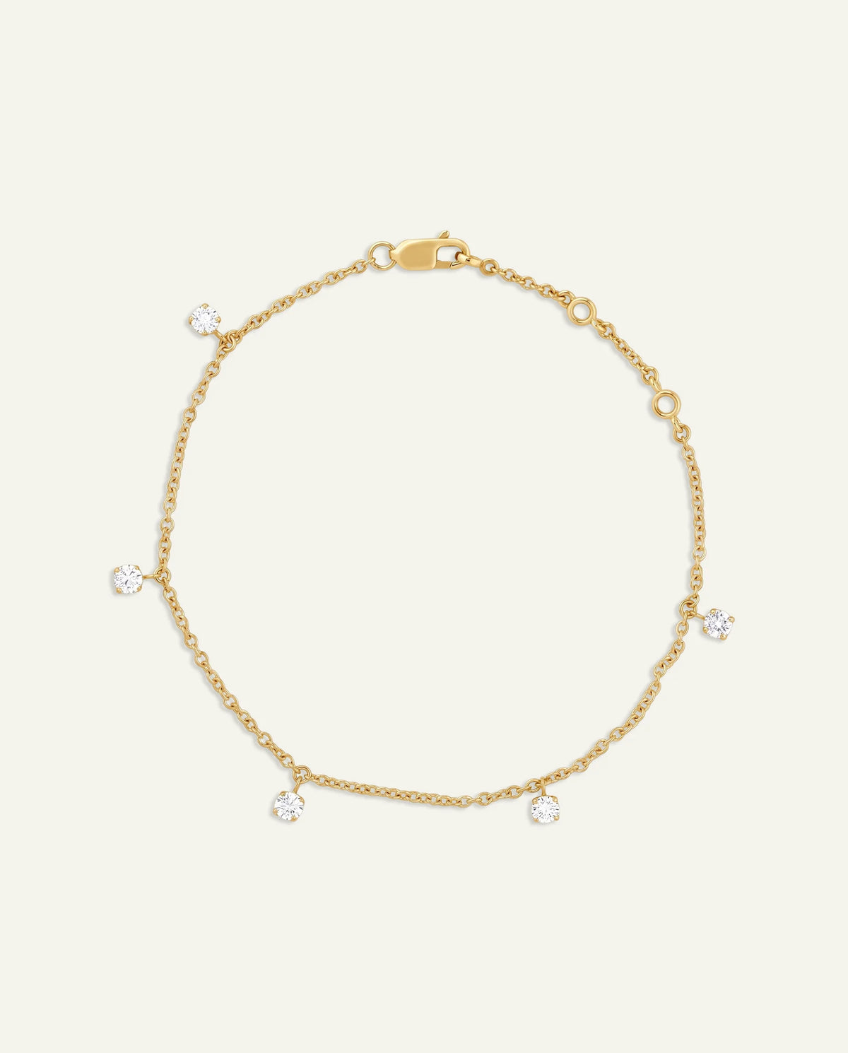 A delicate gold chain anklet with five small, clear round-cut gemstones spaced along the chain, displayed on a light cream background.