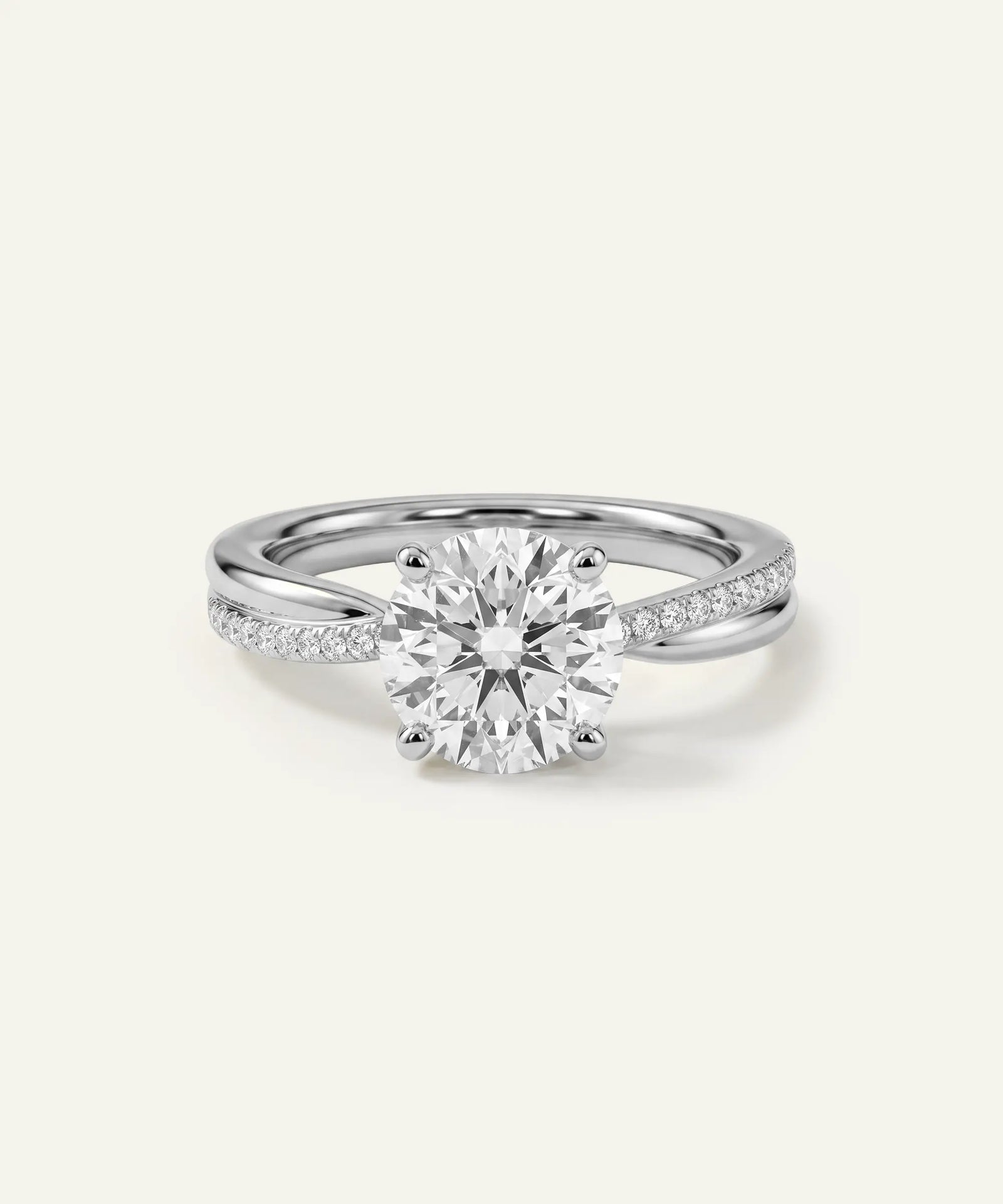 A silver-toned solitaire engagement ring with a round brilliant-cut center stone and pavé-set side stones on a slim band, shown on a light cream background.