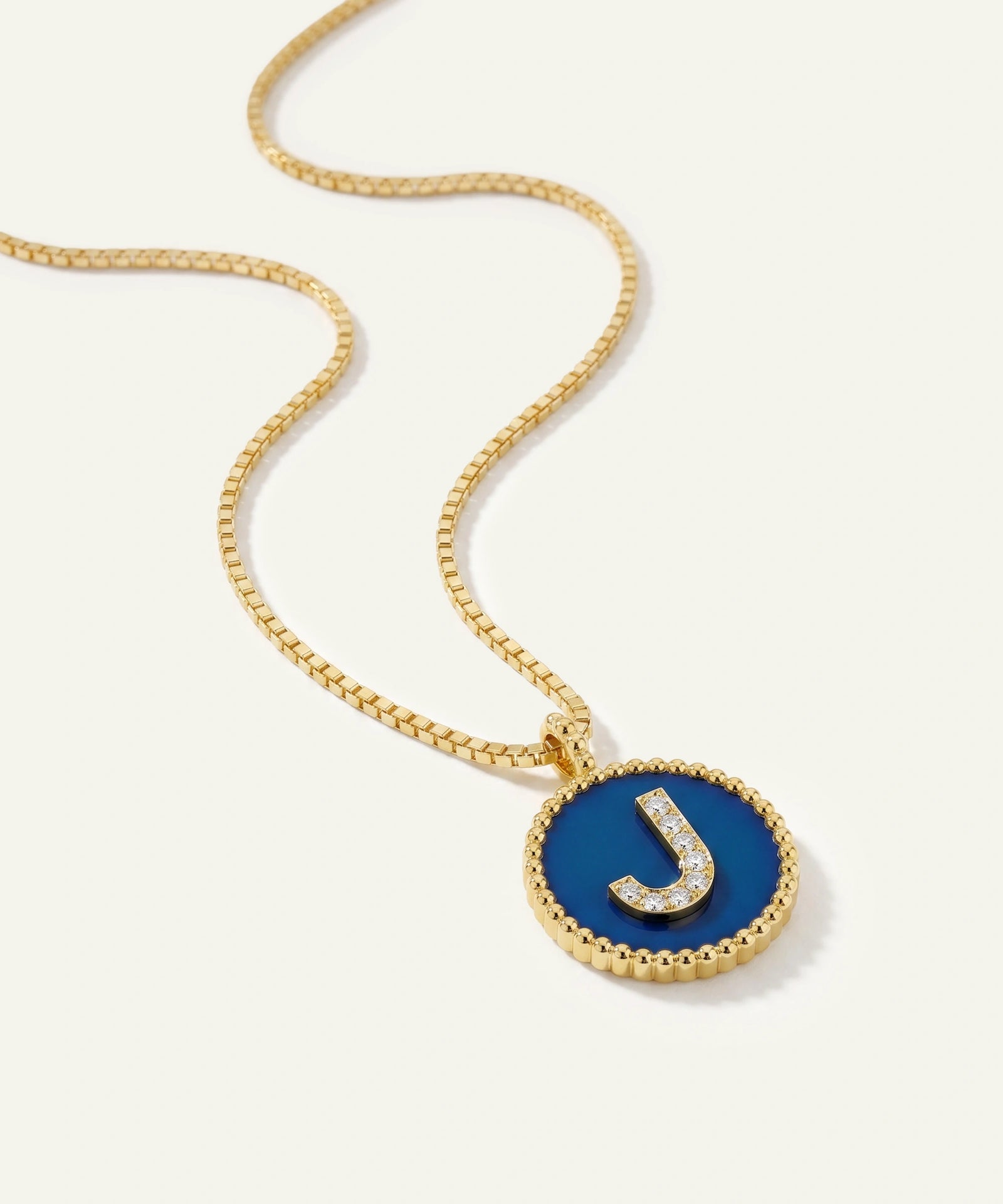 A gold chain necklace with a round bezel pendant featuring a deep blue enamel background and a raised gold letter 'J' accented with small clear stones, shown on a light cream background.