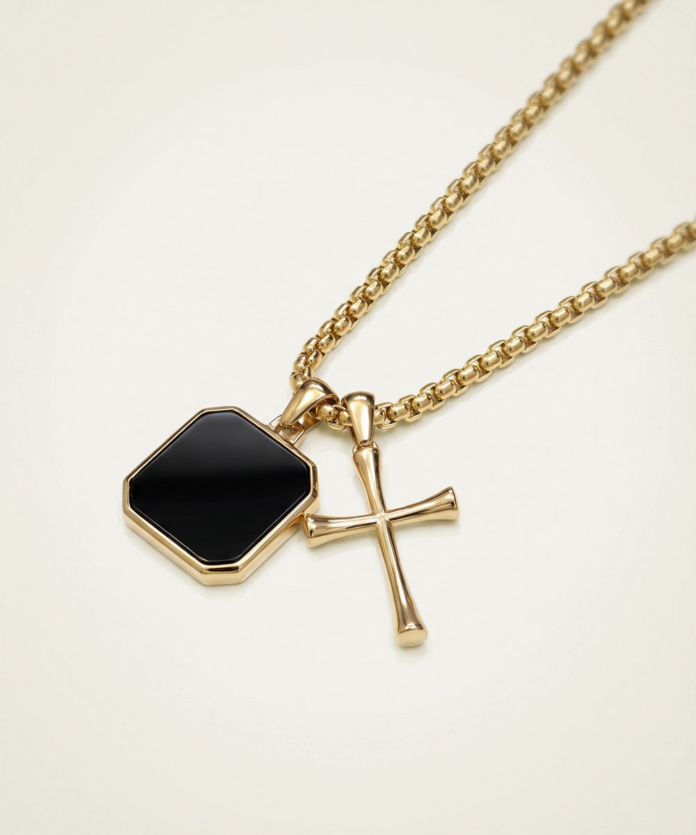 Gold necklace with a black hexagonal pendant and a cross on a beige background