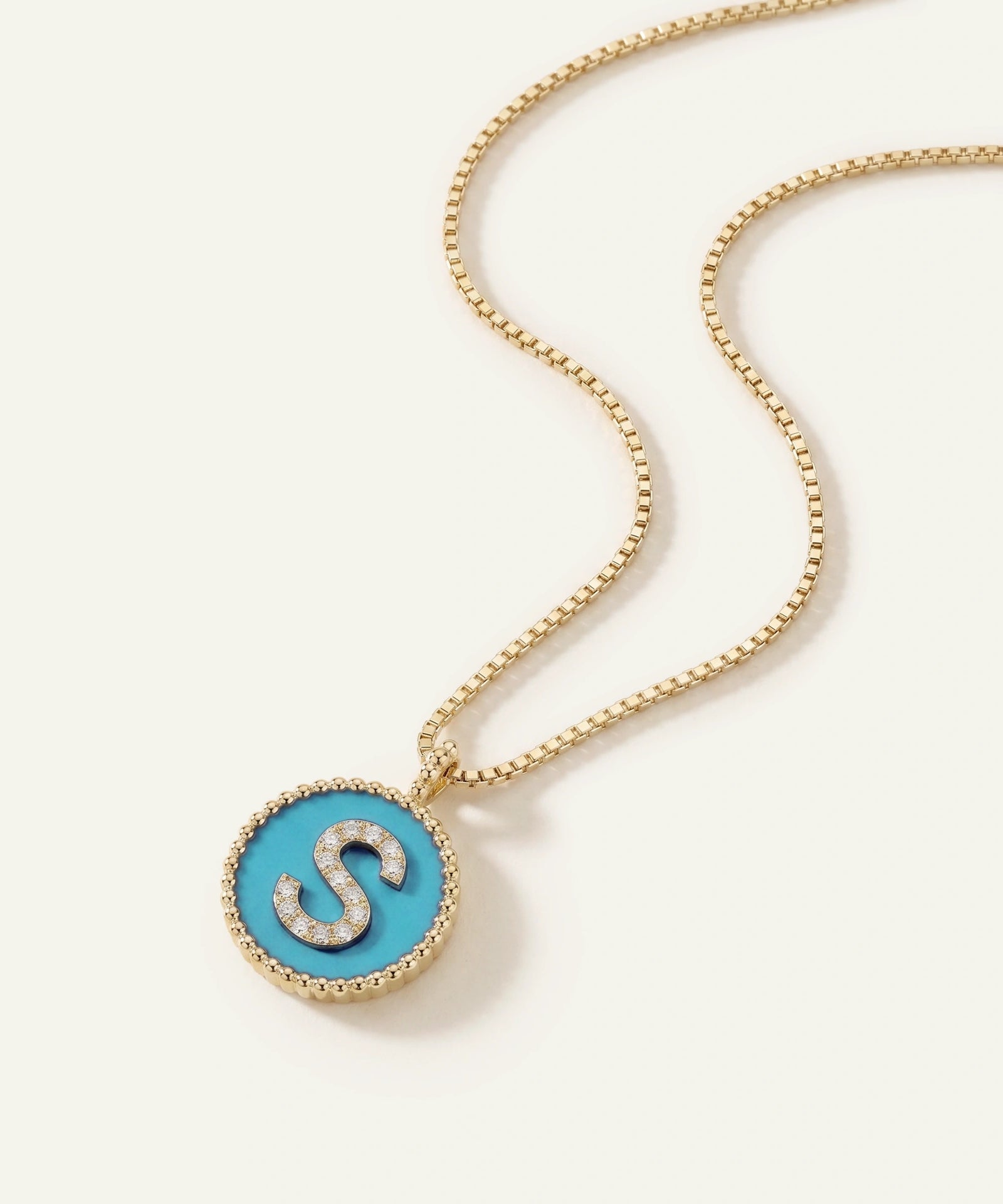 A gold chain necklace with a round pendant featuring a bright turquoise blue center, a silver letter 'S' outlined with small clear stones, and a beaded gold border, shown on a light cream background.