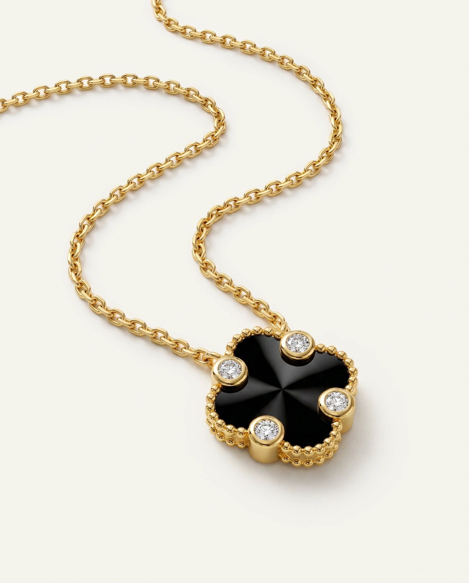 A gold chain necklace featuring a clover-shaped pendant with a glossy black center and four small clear gemstones set around the edges, shown on a light cream background.