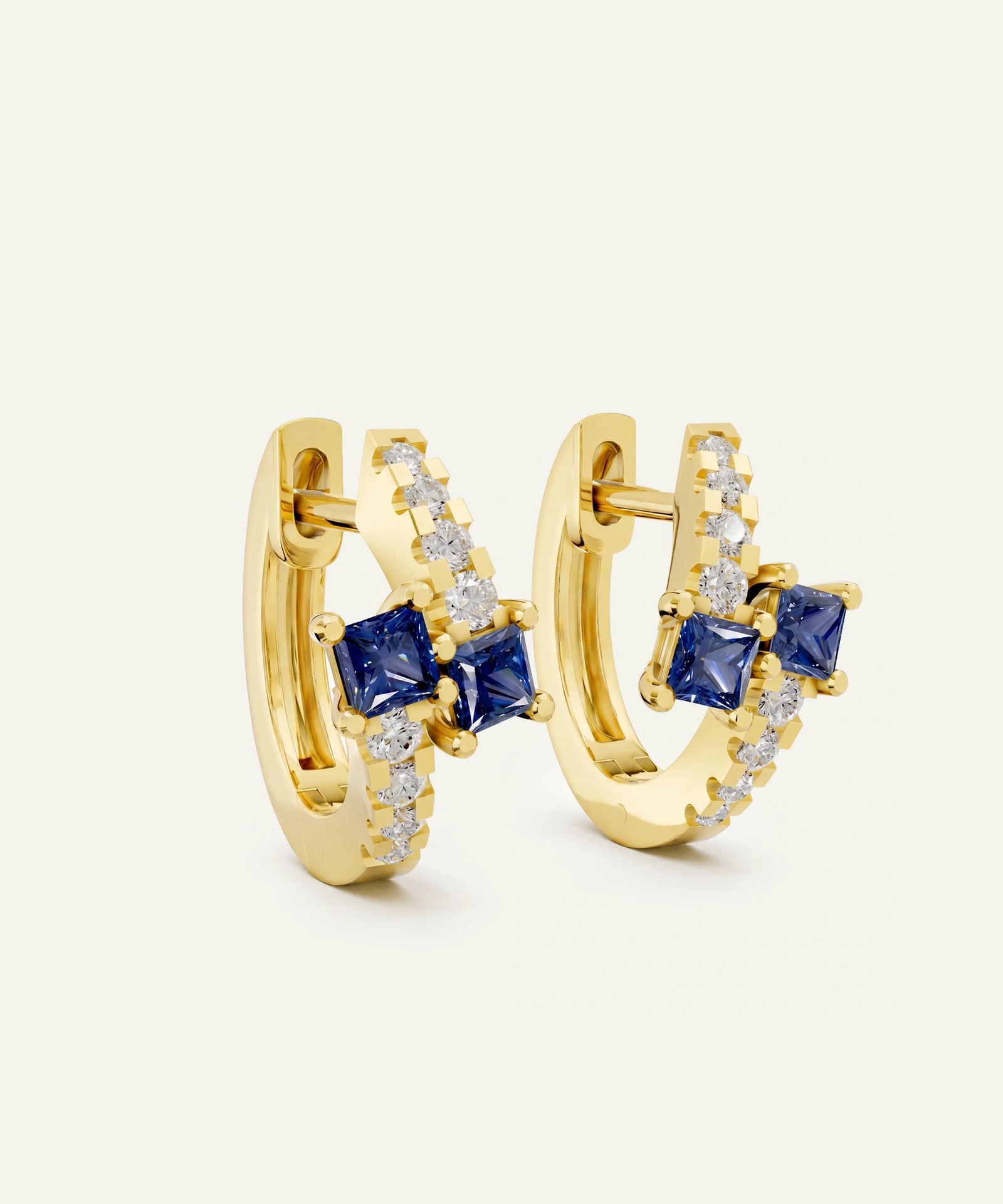 A pair of gold huggie hoop earrings featuring square-cut blue sapphires and rows of small clear stones, set against a solid off-white background.