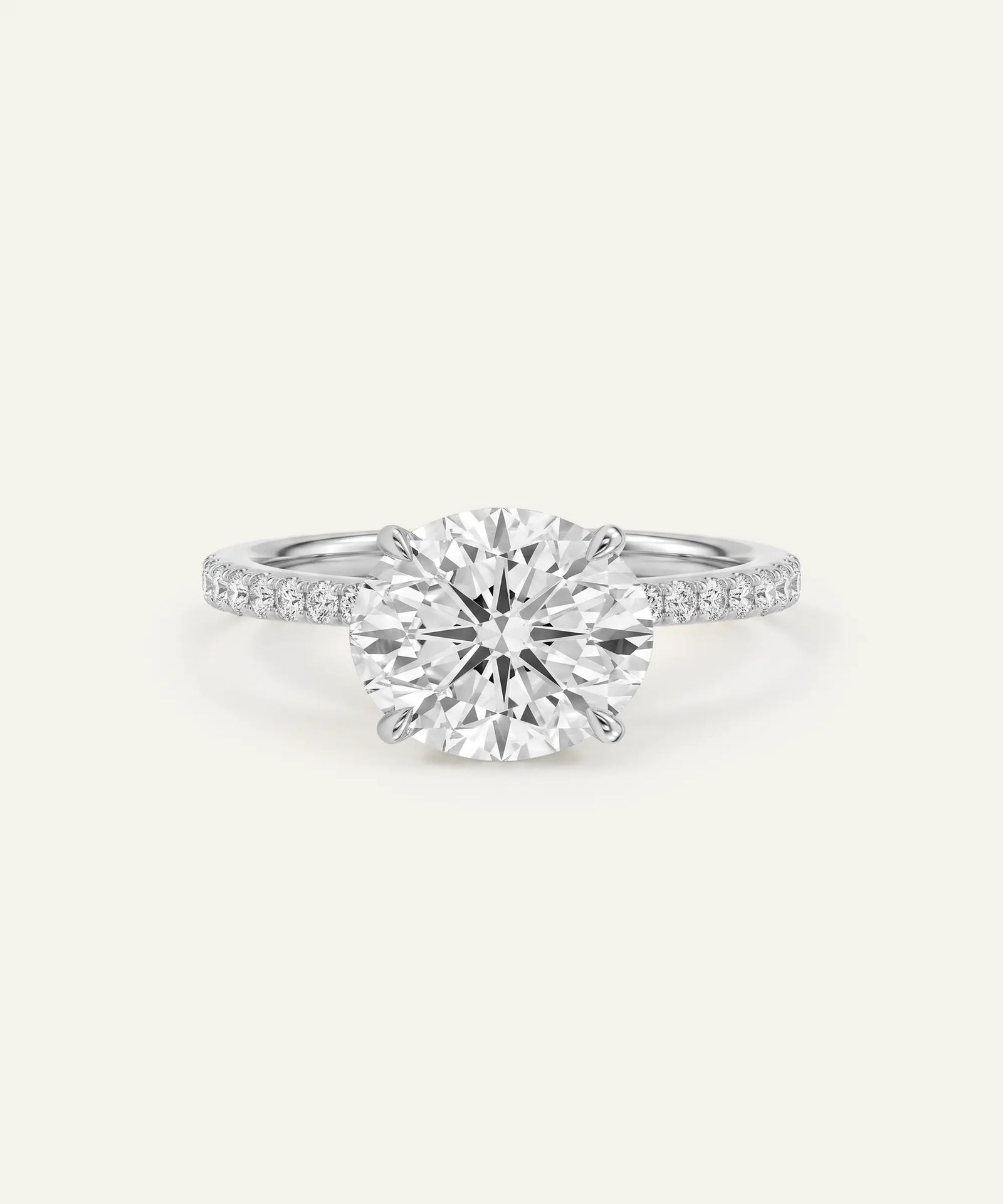 A classic solitaire engagement ring featuring a round brilliant-cut diamond set in a silver-colored metal band with pavé accents on the shoulders, shown against a solid off-white background.