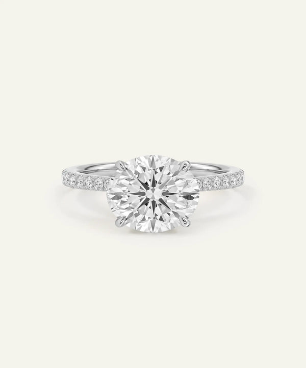 A classic solitaire engagement ring featuring a round brilliant-cut diamond set in a silver-colored metal band with pavé accents on the shoulders, shown against a solid off-white background.