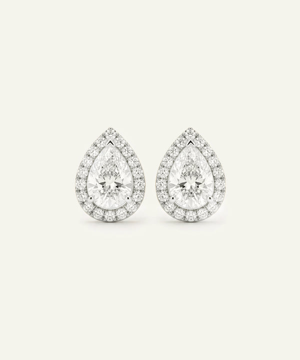 A pair of pear-shaped halo stud earrings in a silver-toned metal, featuring a central round-cut clear gemstone surrounded by a double halo of smaller clear stones, set against a solid off-white background.