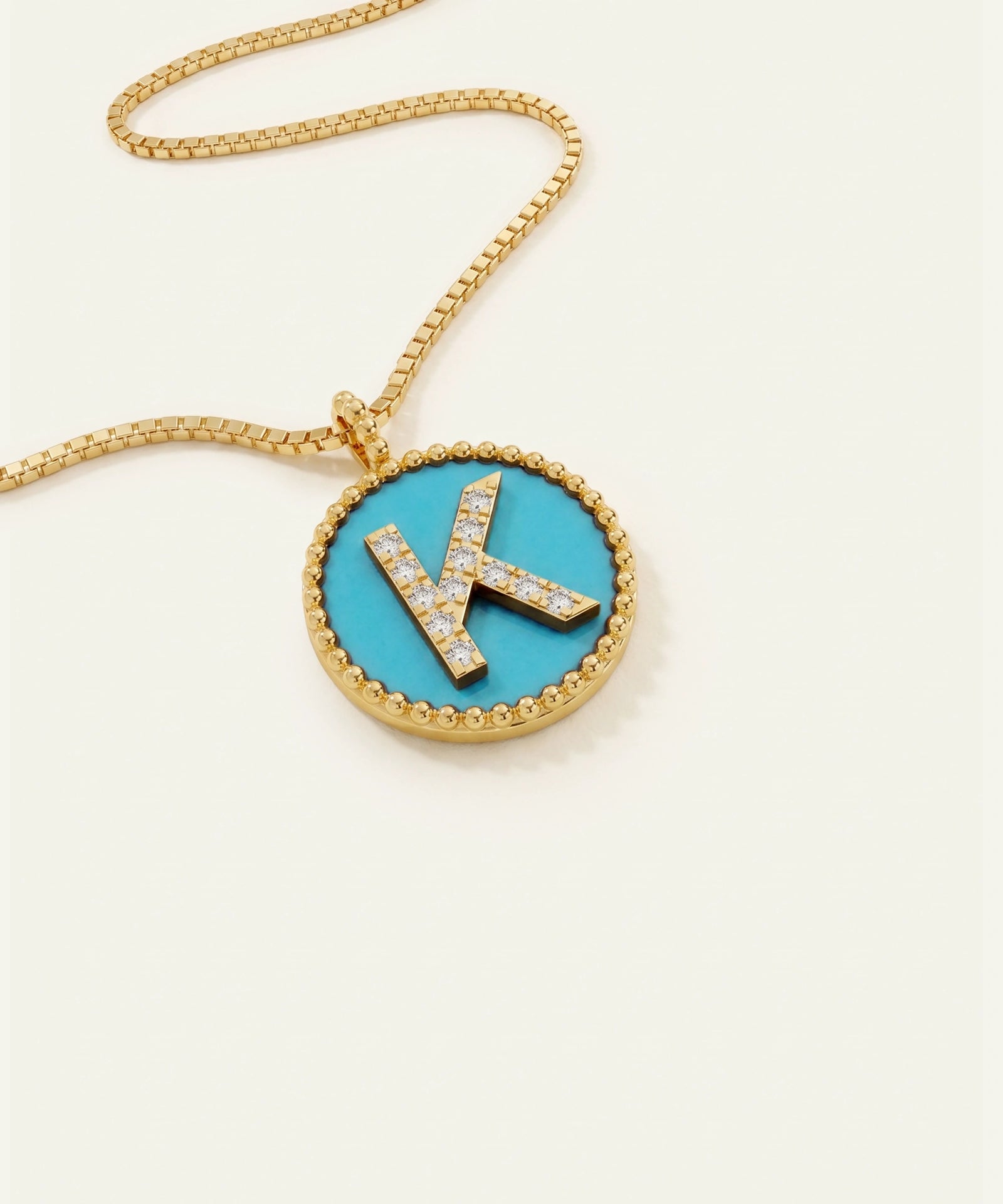 A gold chain necklace with a round bezel pendant featuring a bright turquoise blue background and a raised gold letter 'K' paved with small clear stones, shown on a light cream background.