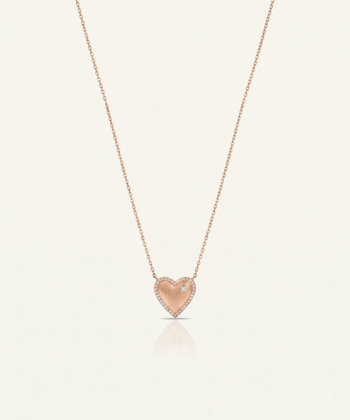 A delicate rose gold heart pendant necklace with a small clear gemstone accent, suspended on a fine chain against a light cream background.