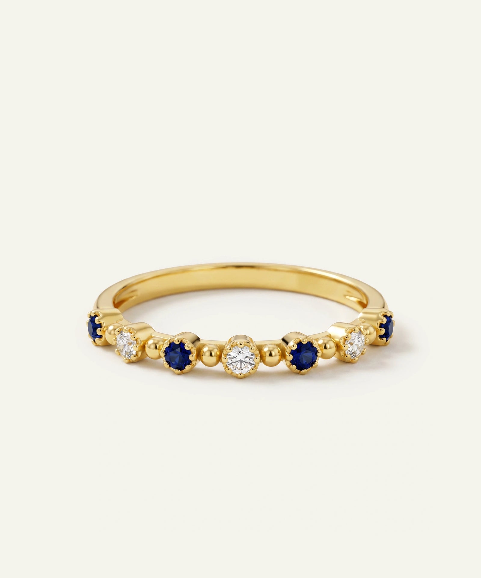 A yellow gold eternity ring featuring alternating round-cut deep blue sapphires and clear round diamonds in a beaded setting, shown on a light cream background.