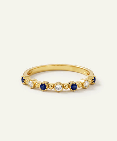 A yellow gold eternity ring featuring alternating round-cut deep blue sapphires and clear round diamonds in a beaded setting, shown on a light cream background.