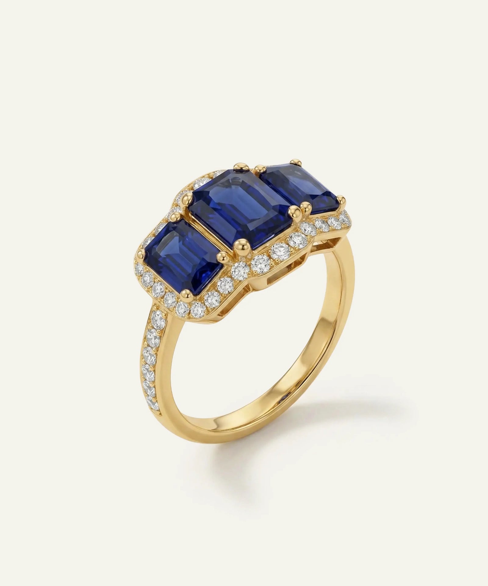 A yellow gold ring featuring three rectangular blue sapphire gemstones in a prong setting, surrounded by small pavé-set clear stones, shown on a light cream background.