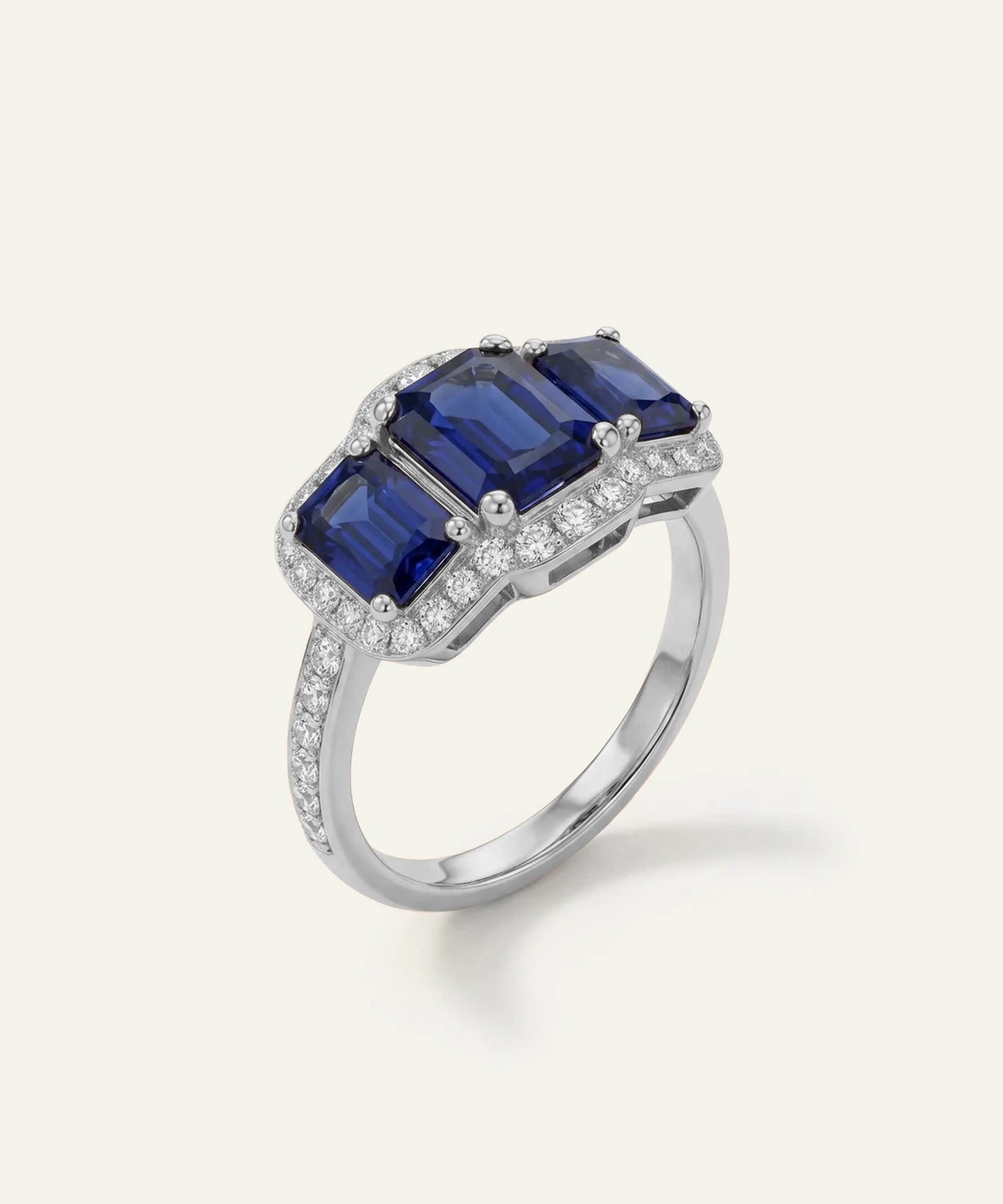 A silver ring featuring three emerald-cut deep blue sapphires in a three-stone cluster setting, surrounded by small pavé-set diamonds, on a light cream background.
