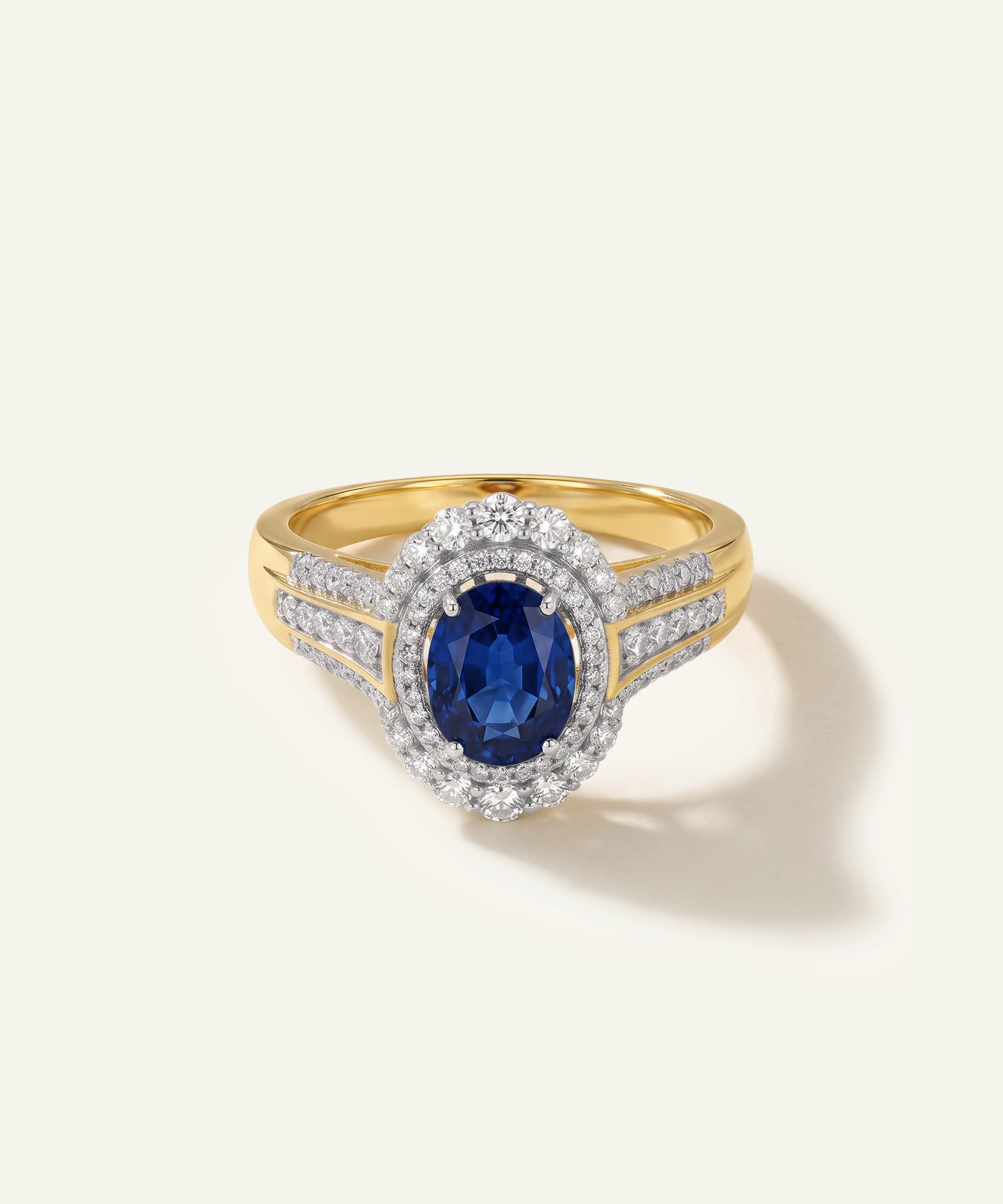 A yellow gold ring featuring an oval-cut blue sapphire center stone surrounded by a halo of small clear stones, with additional clear stones set along the split shank, photographed on a light cream background.