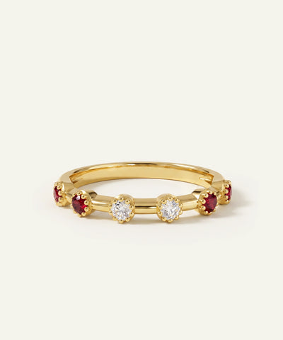 A gold band ring featuring a row of round-cut clear center stones and alternating deep red round side stones, photographed on a clean off-white background.