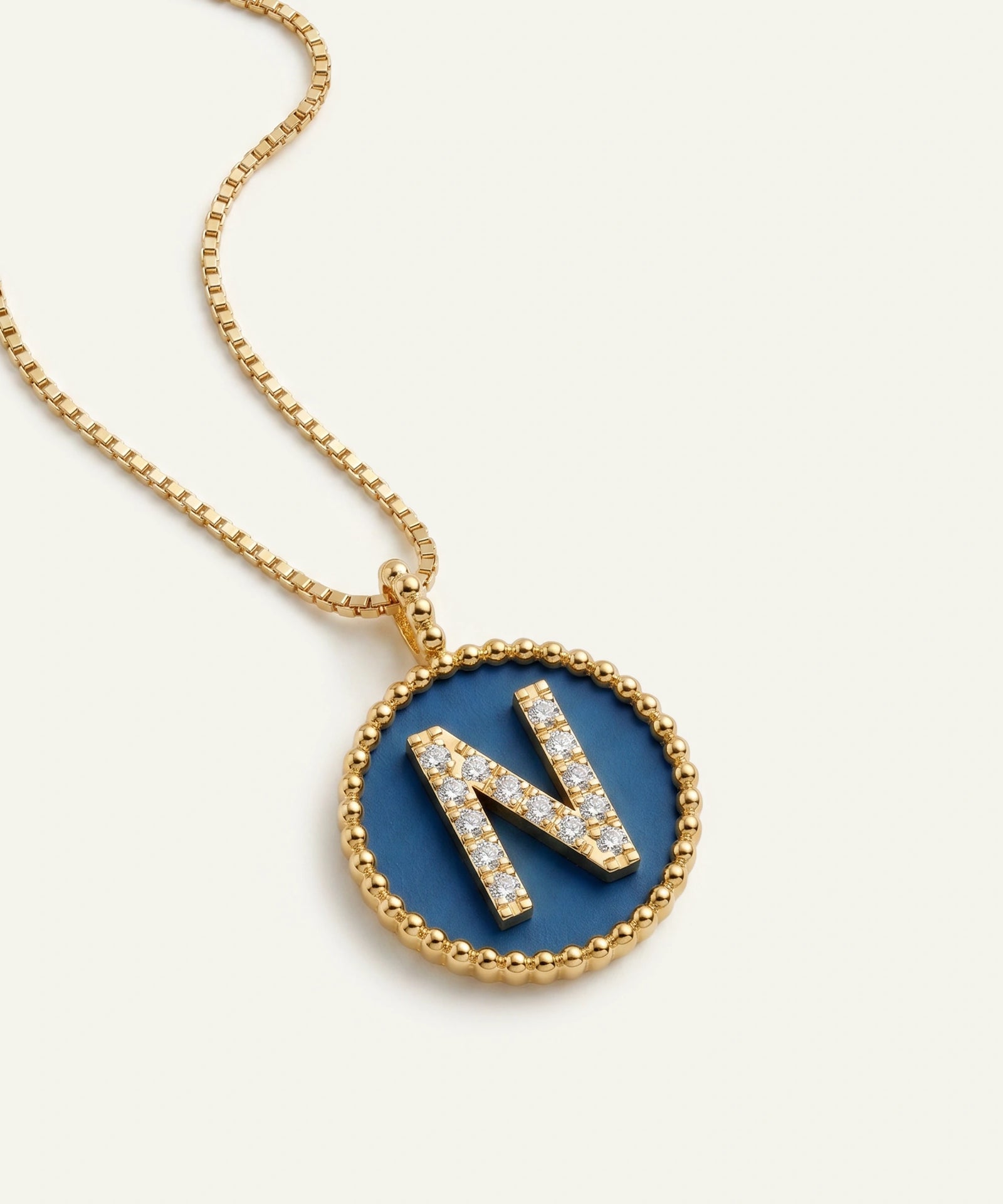 A gold chain necklace with a round bezel pendant featuring a blue enamel background and a raised gold letter 'N' paved with small clear stones, shown on a light cream background.
