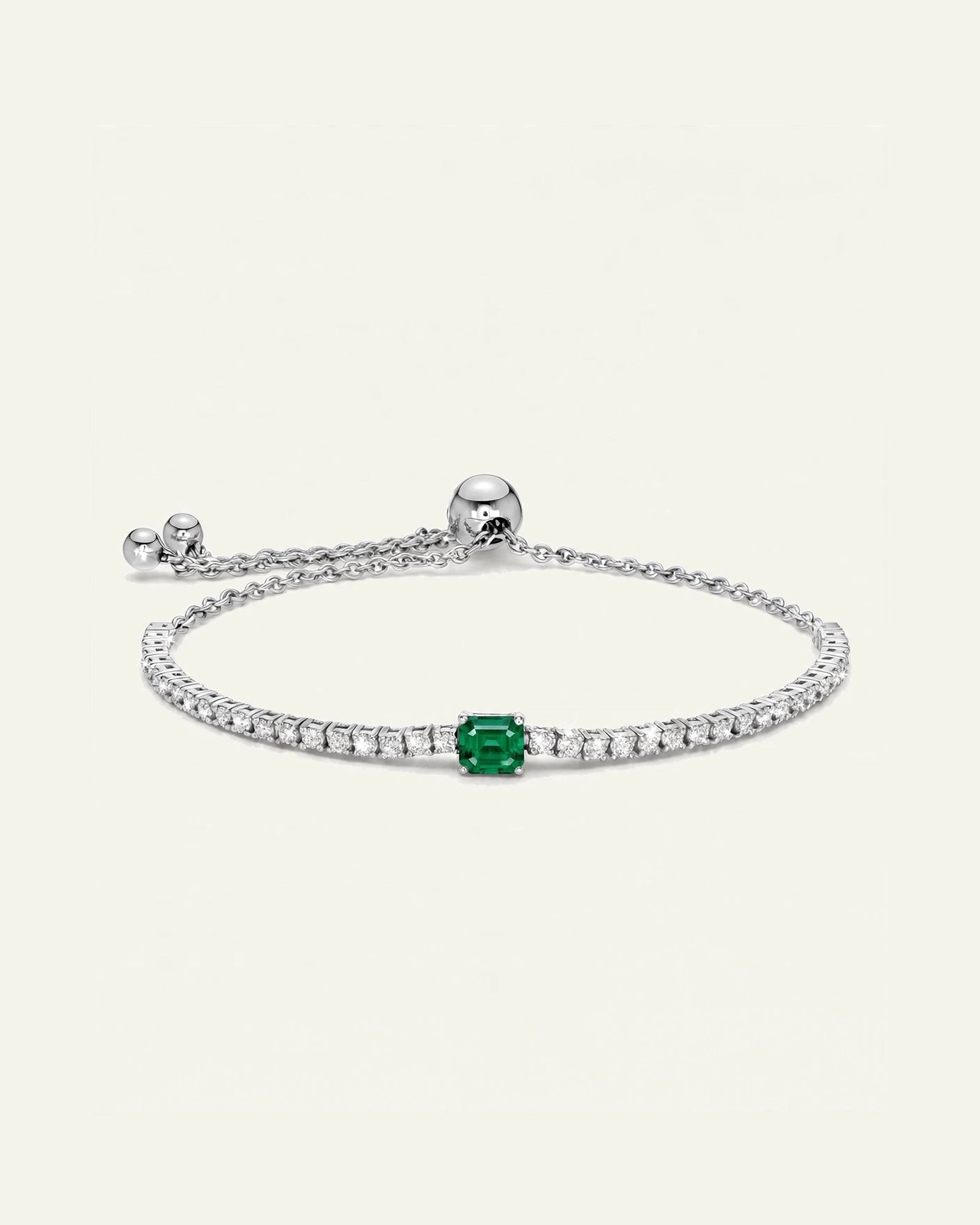 A delicate silver chain bracelet featuring a central emerald-cut green gemstone surrounded by small clear stones, shown on a light cream background.