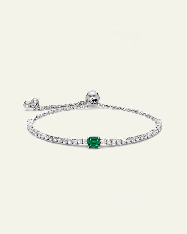 A delicate silver chain bracelet featuring a central emerald-cut green gemstone surrounded by small clear stones, shown on a light cream background.
