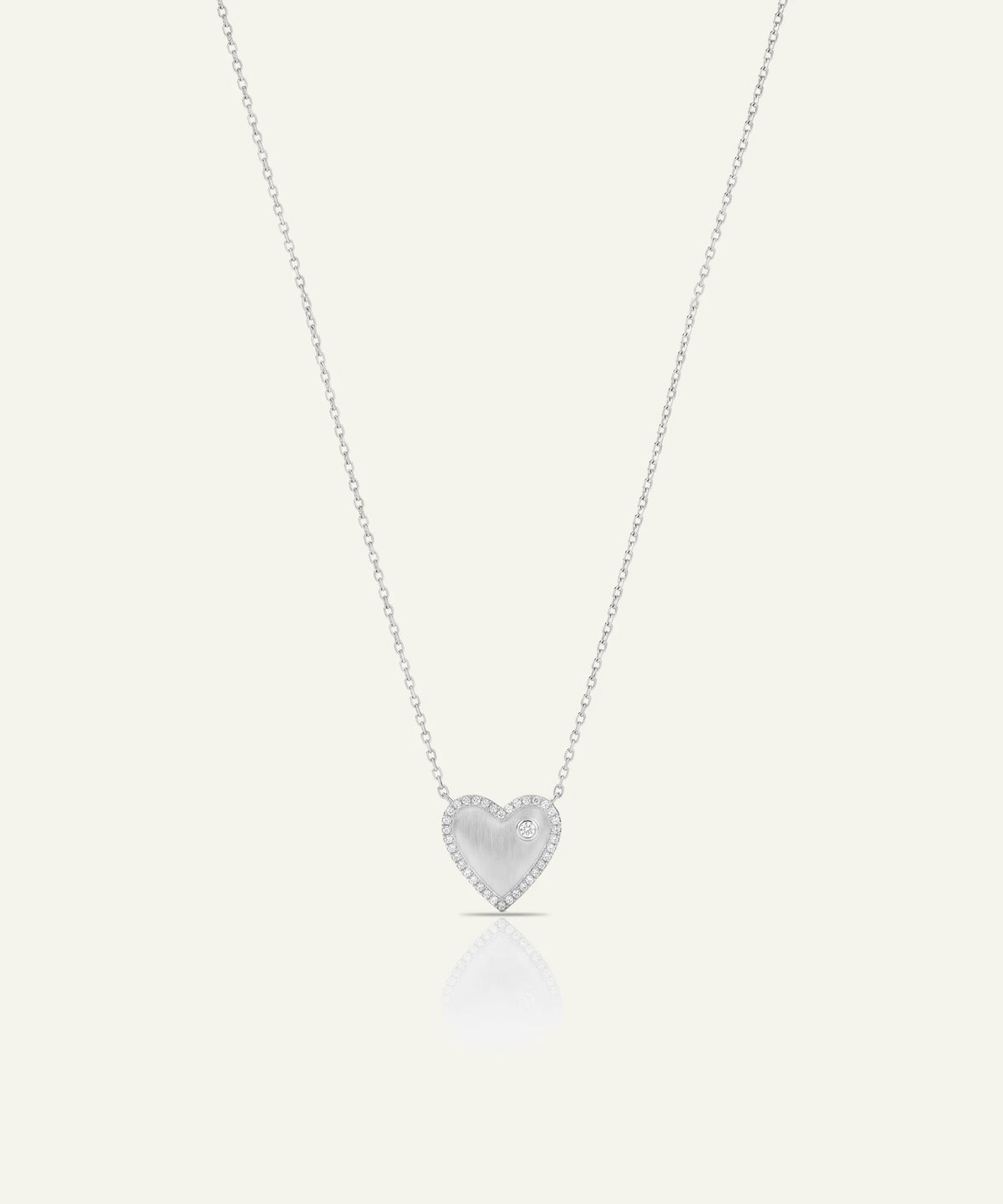 A delicate silver heart pendant necklace with a textured center and a border of small clear stones, suspended on a fine silver chain against a light cream background.