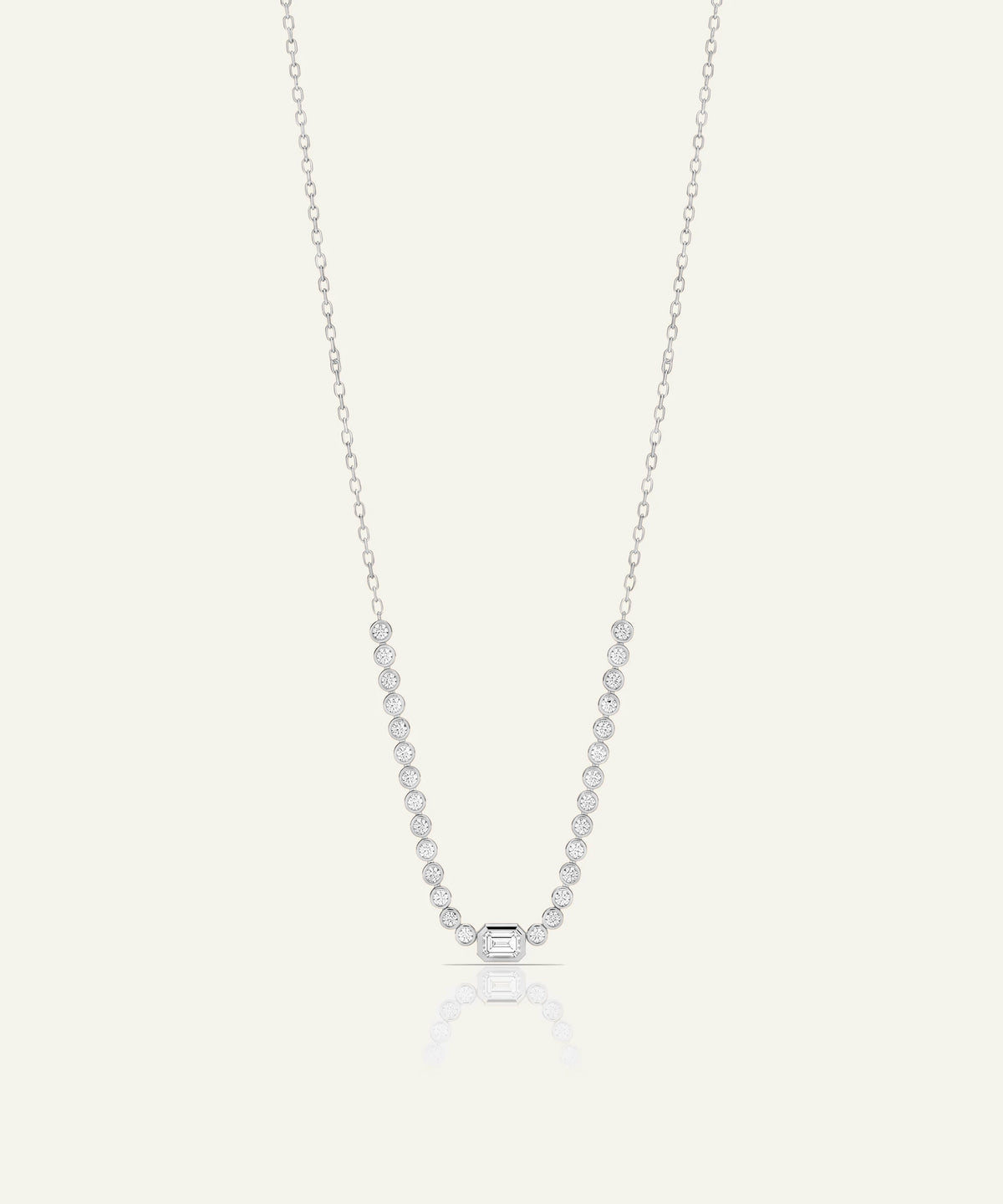 A delicate silver necklace featuring a row of small clear round-cut stones leading to a single larger round stone at the center, displayed against a light cream background.