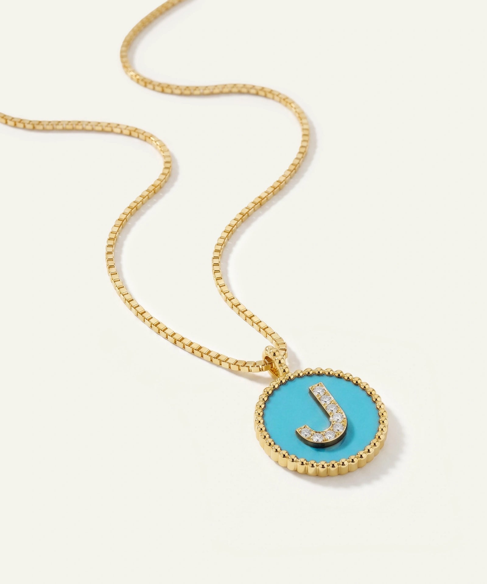 A gold chain necklace with a round bezel pendant featuring a bright turquoise blue center and a raised gold letter 'J' accented with small clear stones, shown on a light cream background.