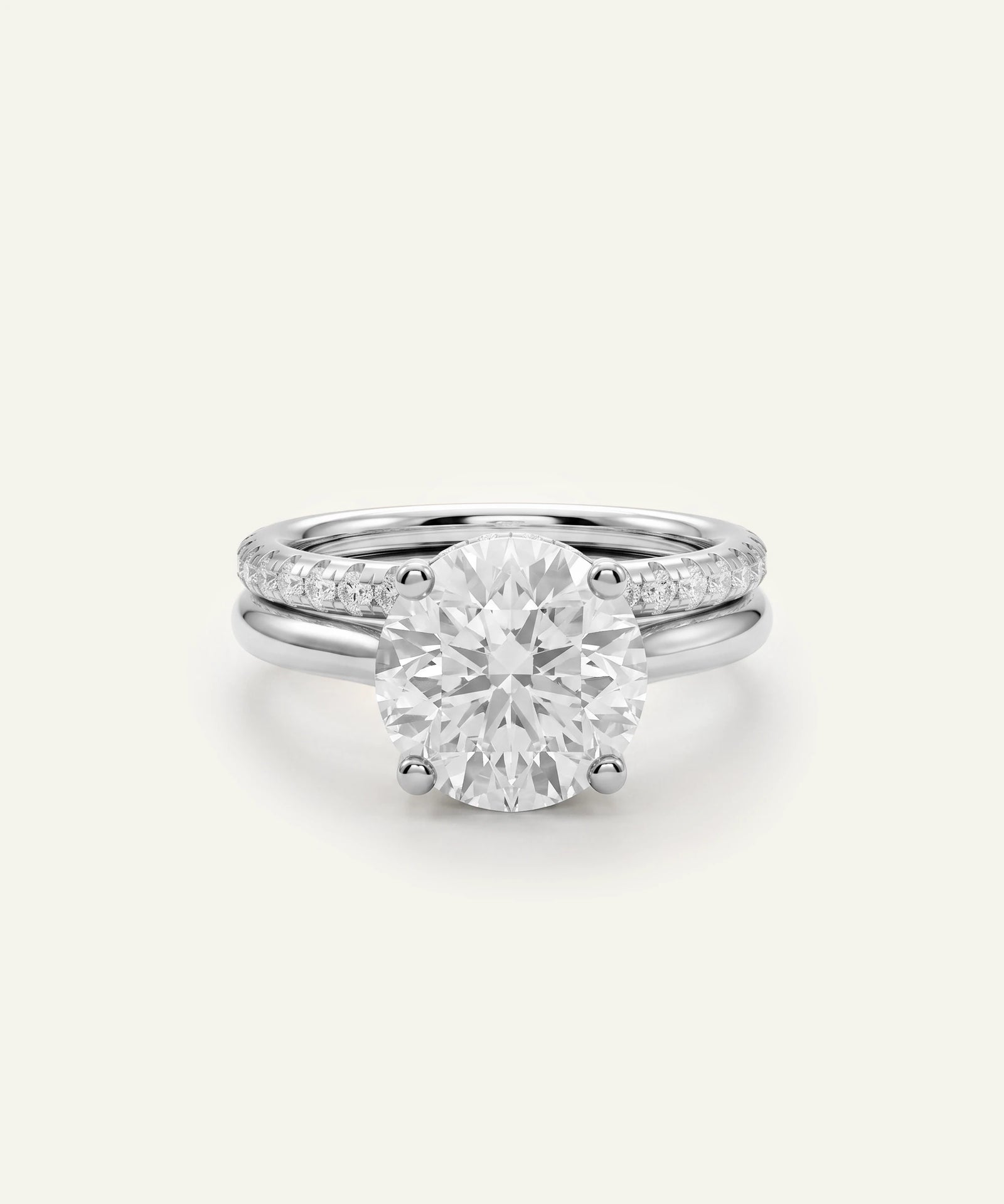 A silver-toned engagement ring featuring a large round brilliant-cut center stone, with a slim pavé band, shown on a light cream background.