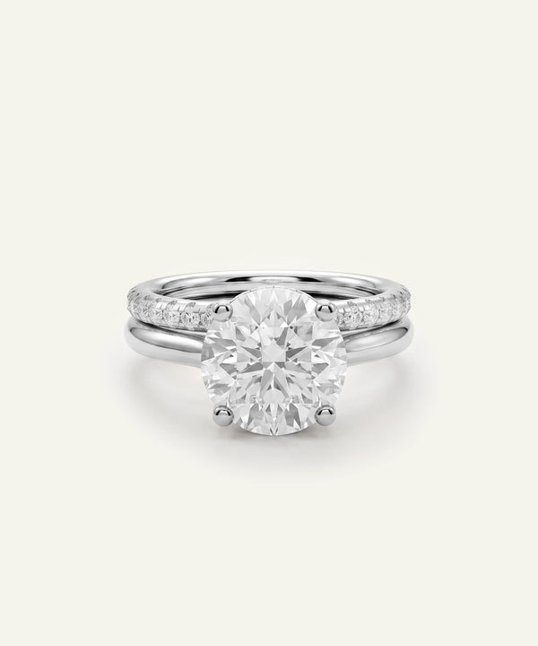 A silver-toned engagement ring featuring a large round brilliant-cut center stone, with a slim pavé band, shown on a light cream background.