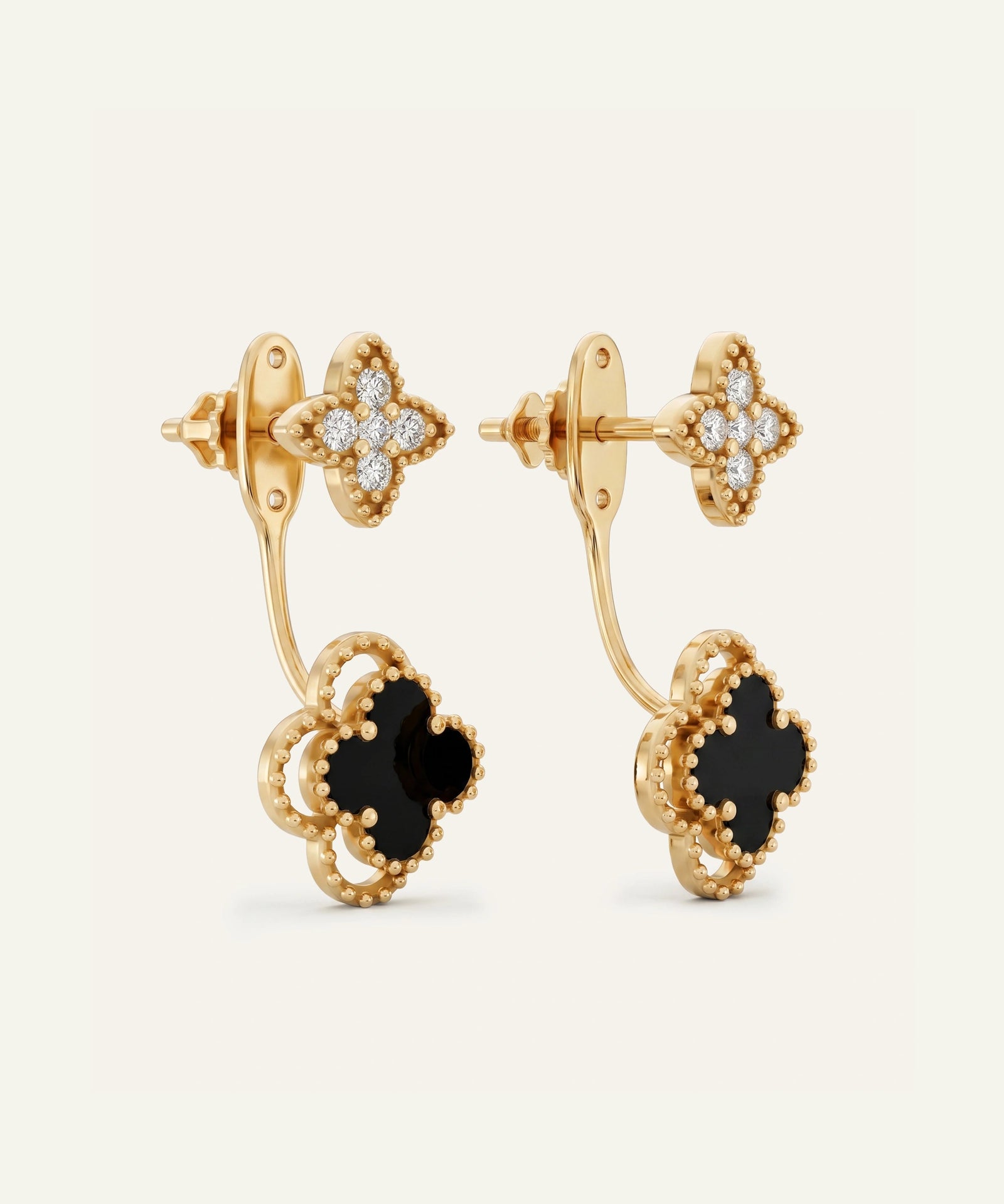 A pair of gold-toned drop earrings with clover-shaped motifs, featuring black onyx-like centers and pavé-set clear stones on the upper loops, shown on a clean off-white background.