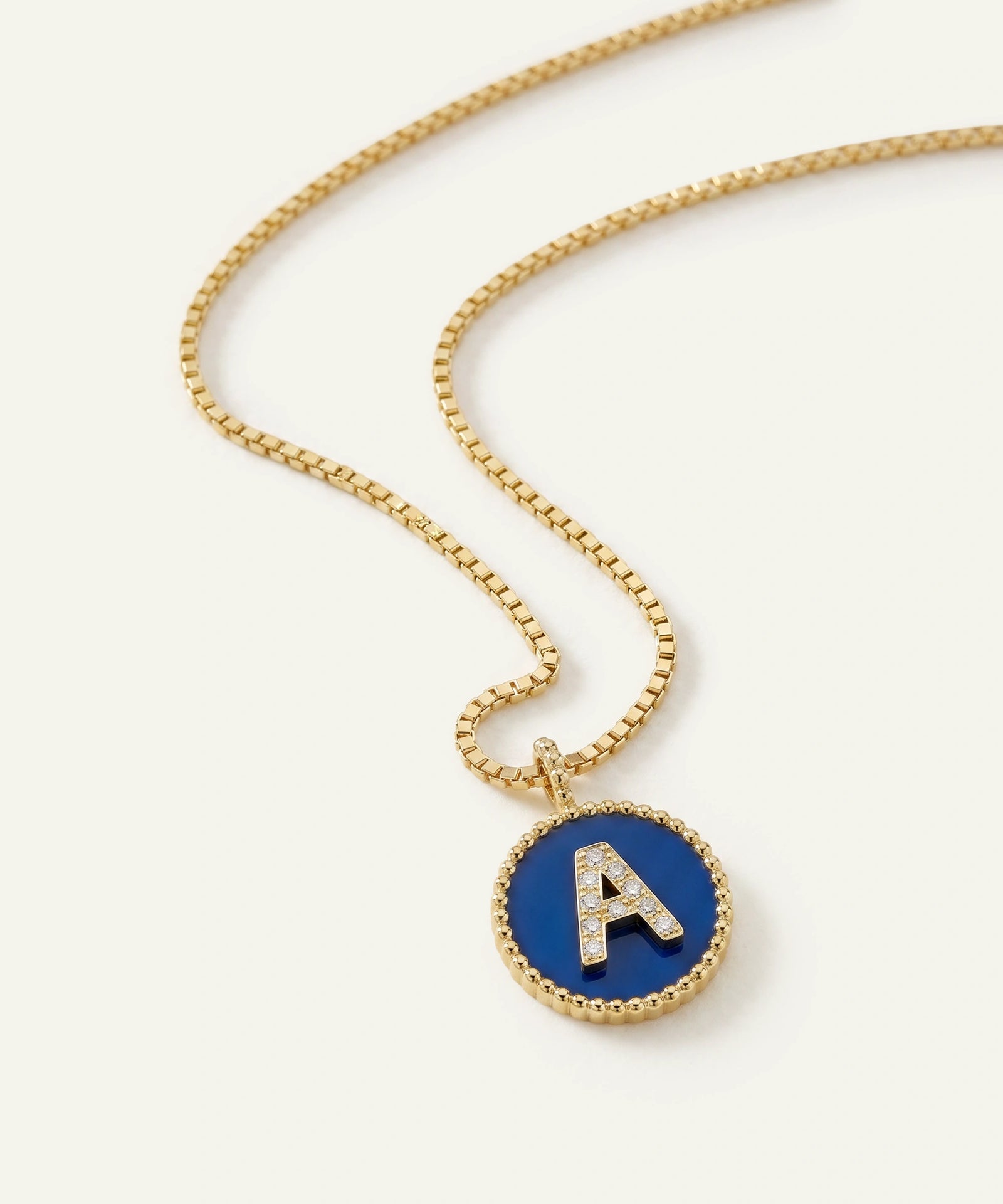 A gold box chain necklace with a circular pendant featuring a deep blue enamel background, a gold beaded border, and a raised gold letter 'A' accented with small clear stones.