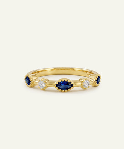 A yellow gold ring set with an oval-cut deep blue sapphire center stone flanked by round clear diamond clusters and smaller blue sapphires, each housed within delicate beaded milgrain bezels, displayed against a solid off-white background.