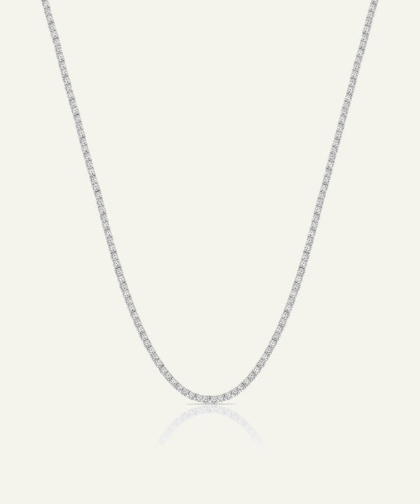 A delicate silver-toned tennis chain necklace with small, clear stones set along the entire chain, displayed against a light cream background with a soft reflection below.