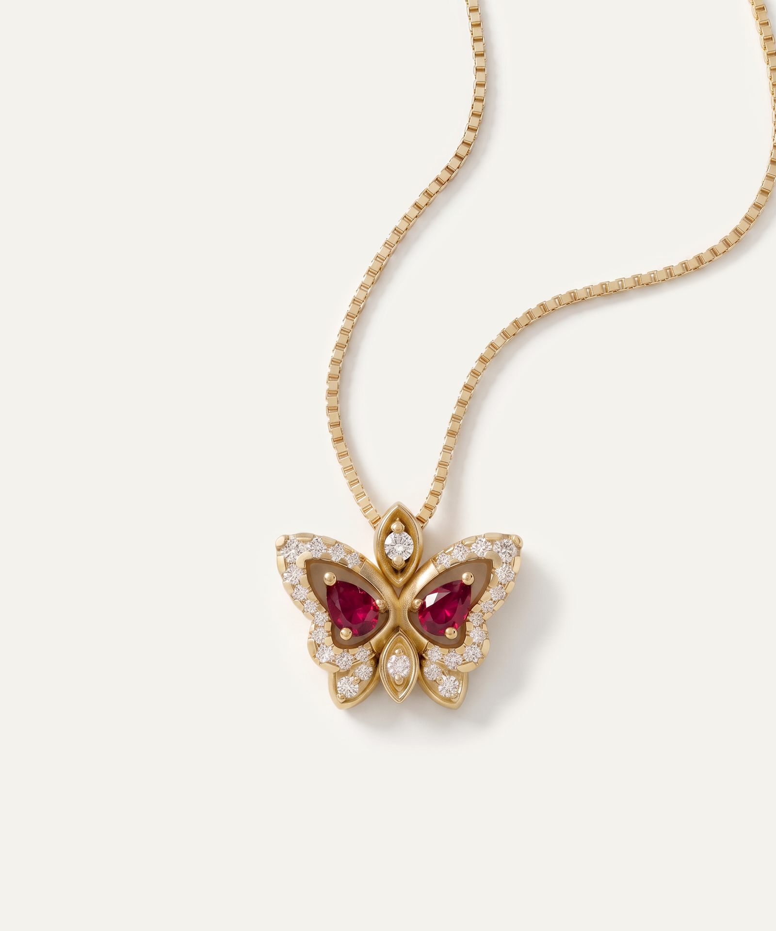 A gold butterfly pendant necklace featuring red gemstone wings and clear pavé-set stones, shown on a clean off-white background.