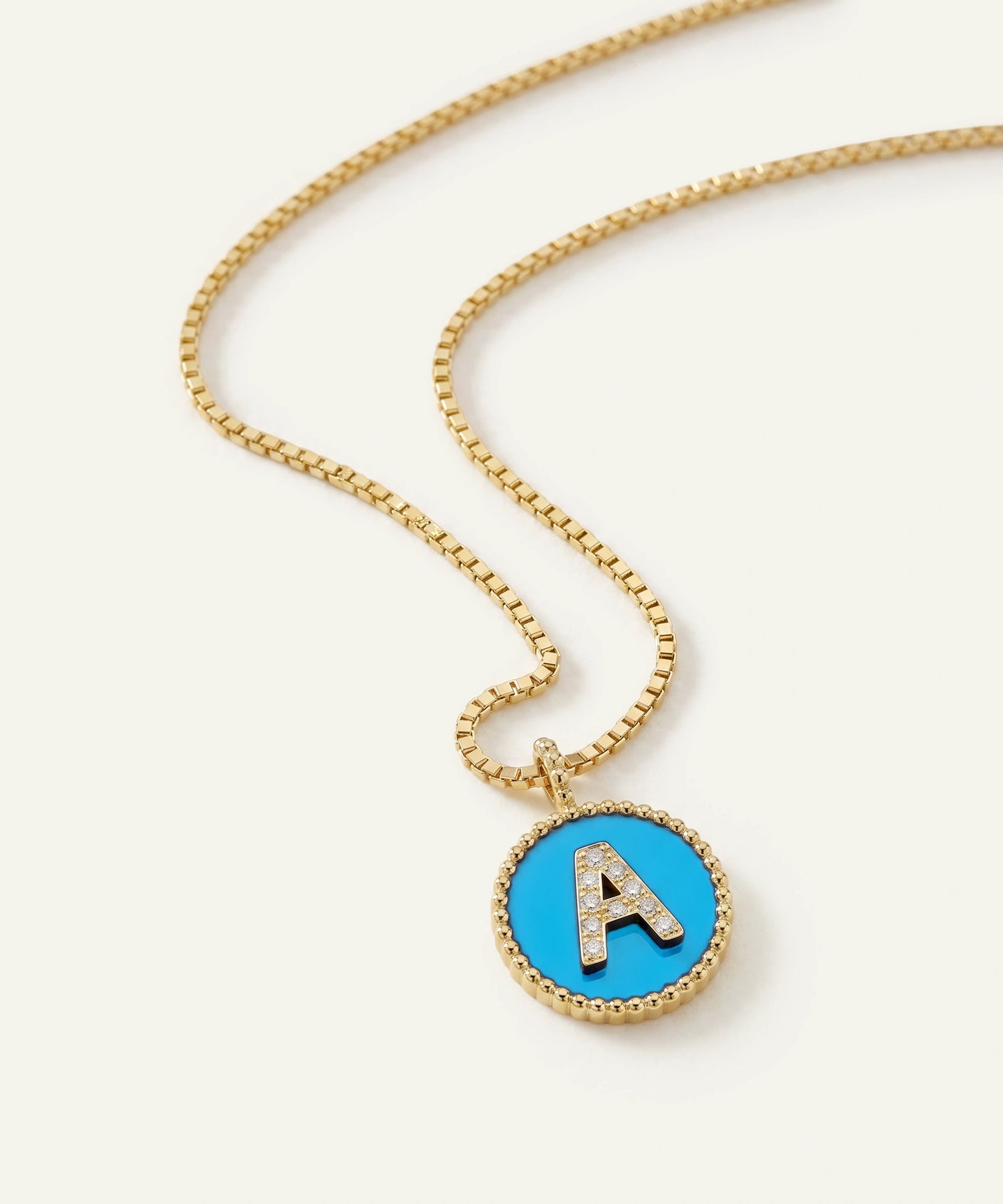 A gold chain necklace with a round pendant featuring a bright blue center, a gold beaded border, and a raised gold letter 'A' embellished with small clear stones, shown on a light cream background.