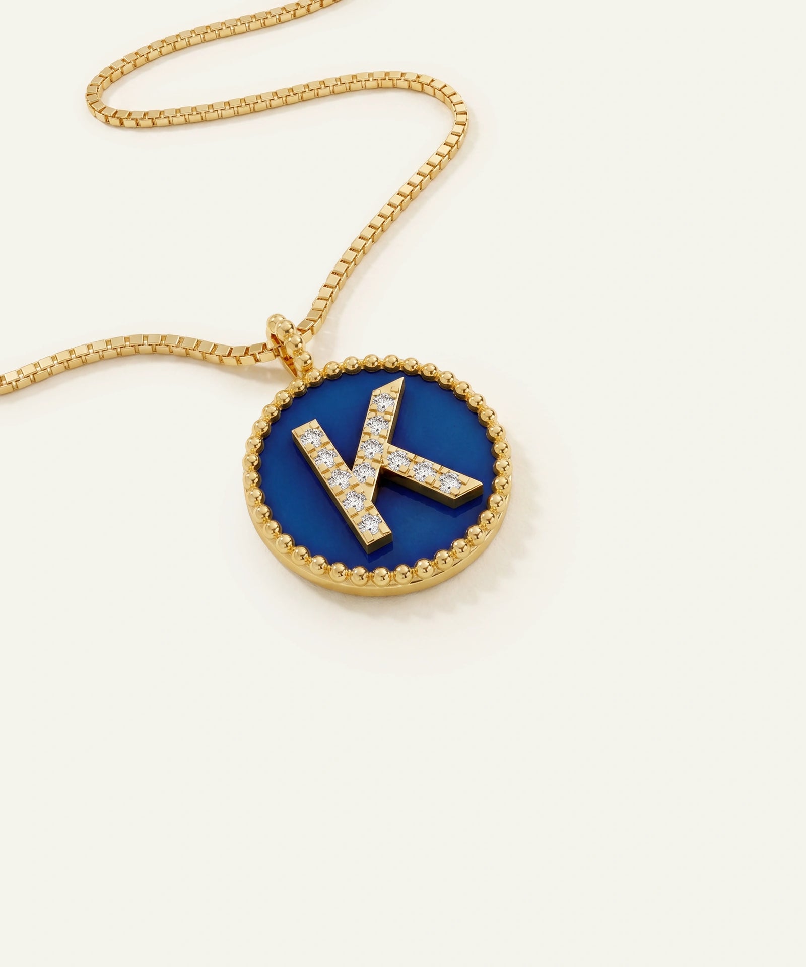 A gold chain necklace with a round pendant featuring a deep blue enamel background and a raised gold letter 'K' paved with small clear stones, shown on a light cream background.